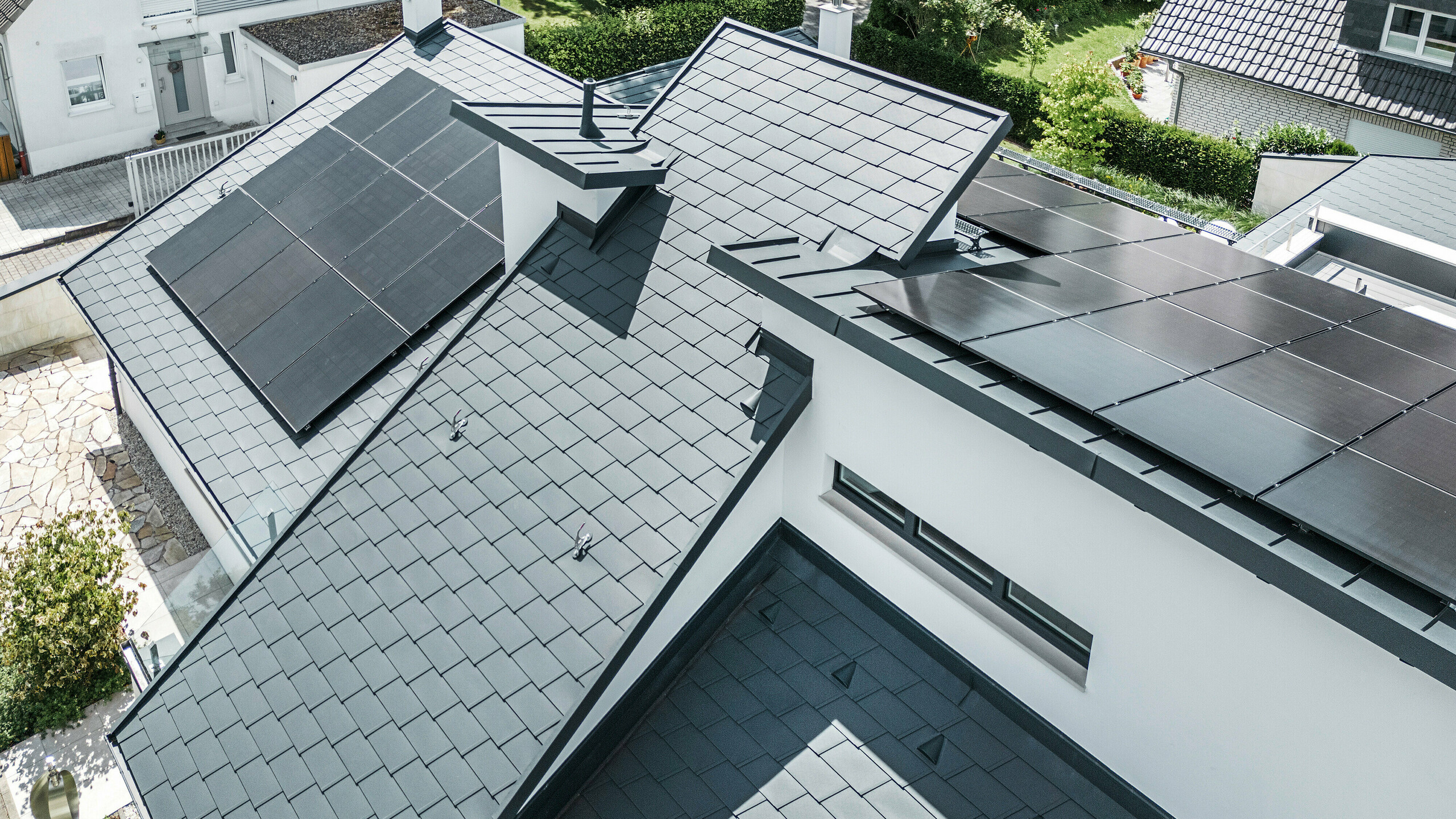 Pohľad z vtáčej perspektívy na rôzne úrovne strechy moderného rodinného domu v Dortmunde, na ktorom boli použité strešné šindle PREFA DS.19 a PREFALZ vo farbe P.10 antracit, ako aj veľkoplošný fotovoltický systém.