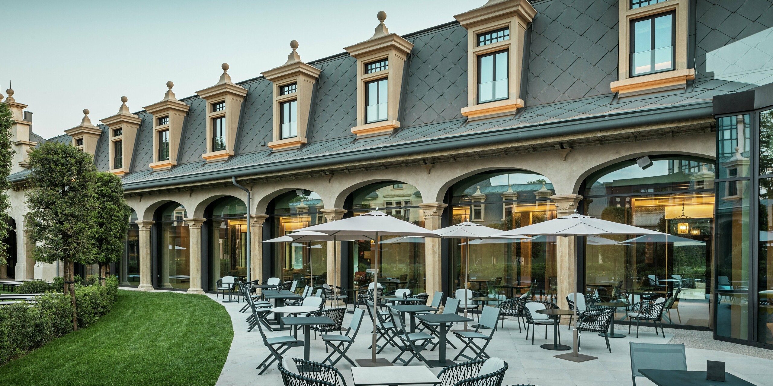 Štýlovo navrhnutá terasa termálneho parku „De Montel – Terme Milano“ láka na oddych vďaka moderným sedeniam a slnečníkom. V pozadí sa stretáva pamiatkovo chránená architektúra s modernou funkčnosťou: hliníková strecha s PREFA strešnými šablónami 44 × 44 v odtieni P.10 svetlošedá harmonicky zapadá do jemne zrekonštruovanej secesnej fasády. Rozsiahle sklenené fasády vytvárajú transparentnosť a elegantným spôsobom spájajú vnútorný a vonkajší priestor.