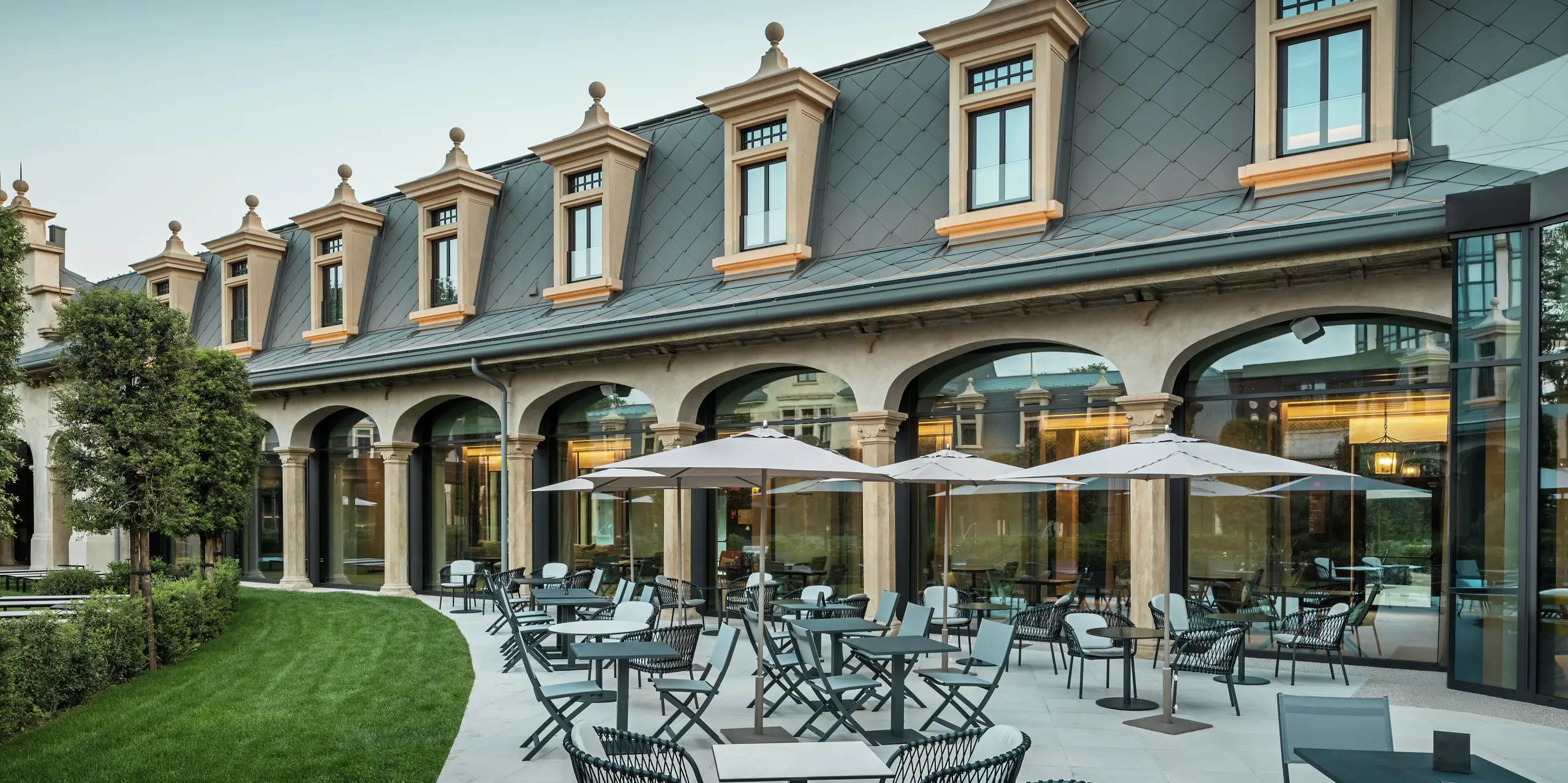 Štýlovo navrhnutá terasa termálneho parku „De Montel – Terme Milano“ láka na oddych vďaka moderným sedeniam a slnečníkom. V pozadí sa stretáva pamiatkovo chránená architektúra s modernou funkčnosťou: hliníková strecha s PREFA strešnými šablónami 44 × 44 v odtieni P.10 svetlošedá harmonicky zapadá do jemne zrekonštruovanej secesnej fasády. Rozsiahle sklenené fasády vytvárajú transparentnosť a elegantným spôsobom spájajú vnútorný a vonkajší priestor.