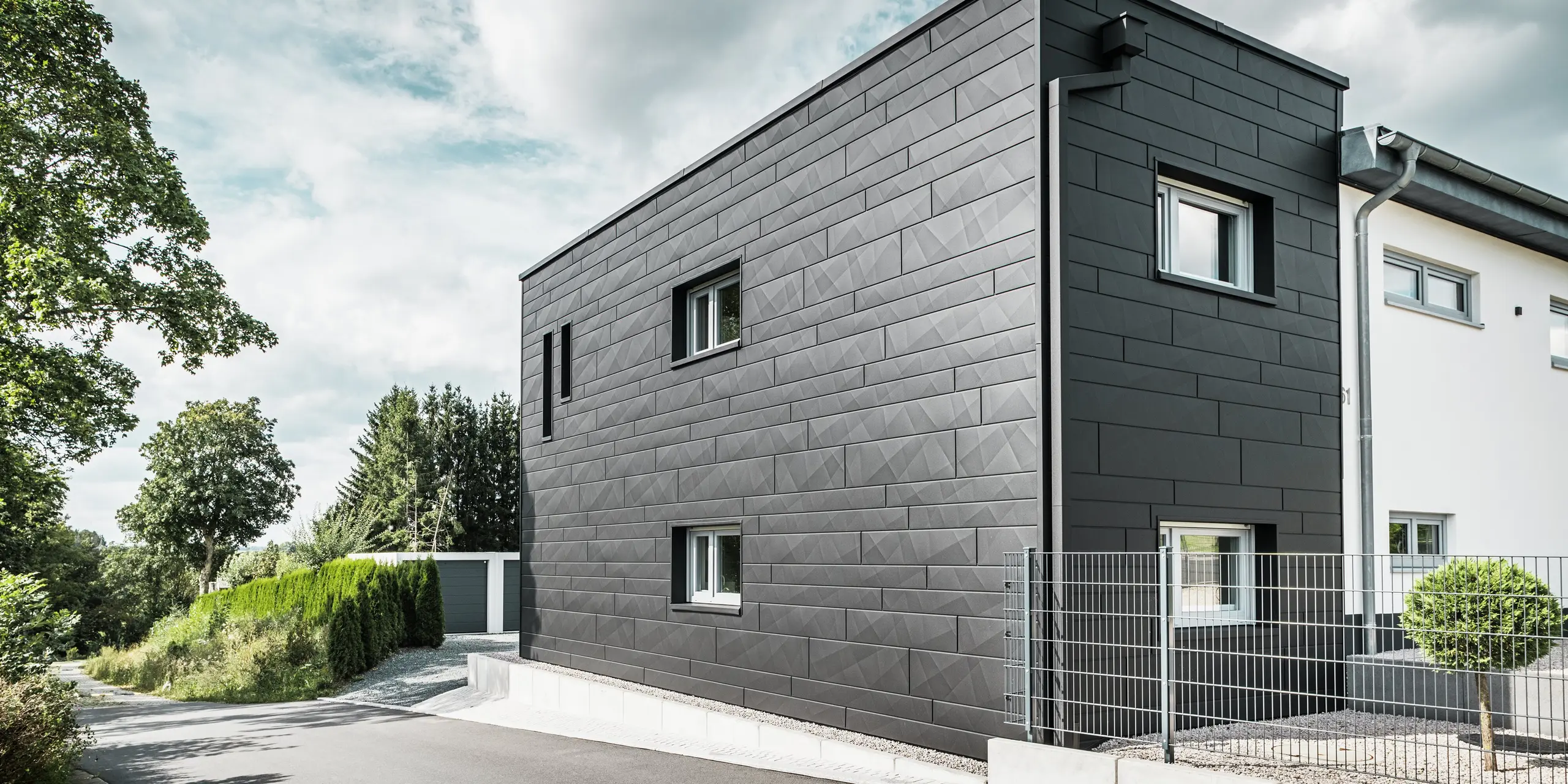 Zadná časť zmodernizovaného rodinného domu v Teuschnitzi zaujme elegantnou fasádou z PREFA Siding.X v čiernej farbe P.10. Geometrické plechové panely vytvárajú výrazný, štruktúrovaný vzhľad a dodávajú budove moderný a nadčasový charakter. Integrovaný odvodňovací systém PREFA s hranatými zvodmi a zberným kotlíkom na vodu sa harmonicky začleňuje do celkového vzhľadu a spája estetiku s funkčnosťou.