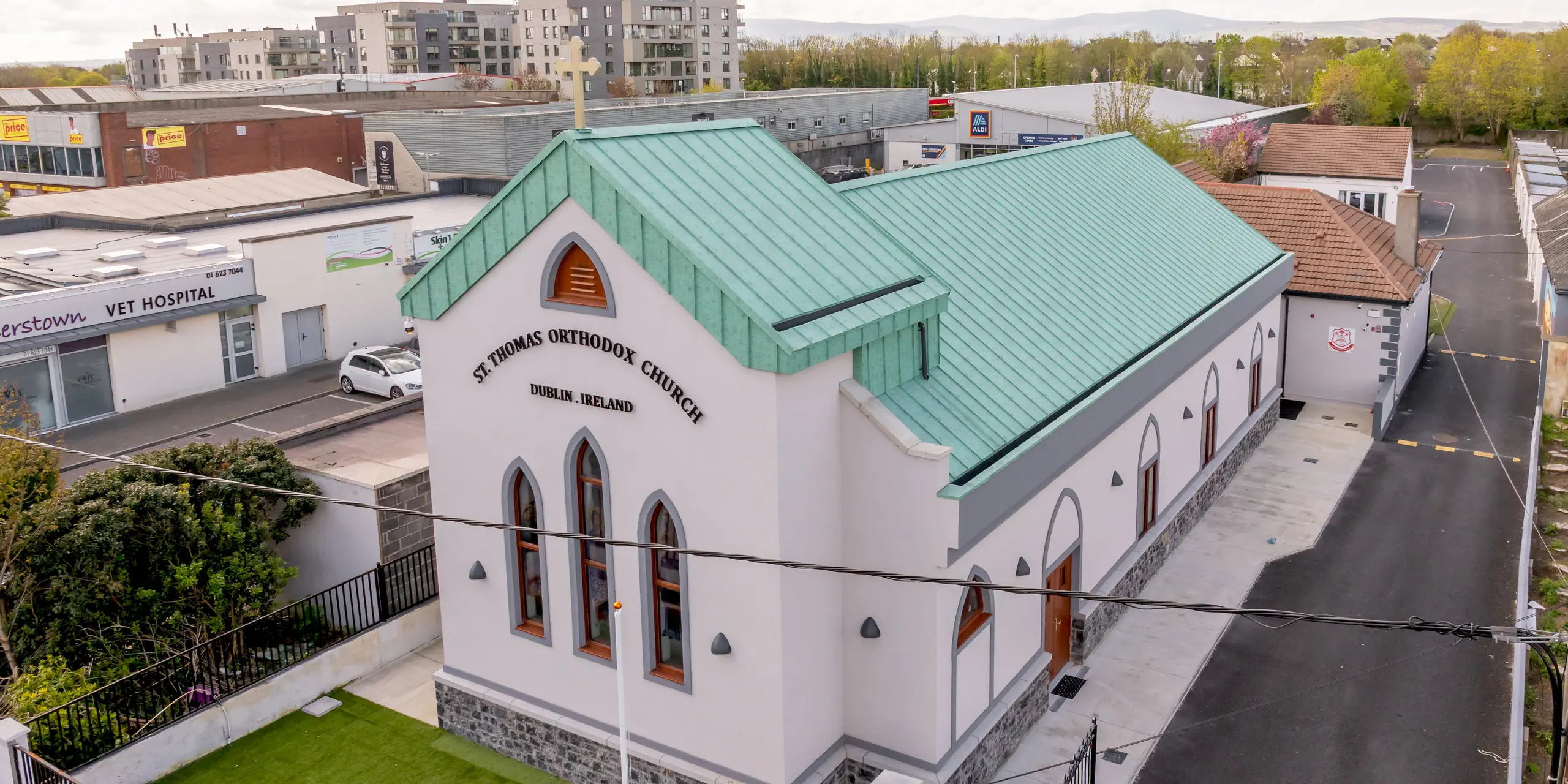 Drohnenaufnahme der St. Thomas Orthodox Church in Palmersgate, Dublin, mit ihrem markanten PREFALZ Aluminium-Stehfalzdach in P.10 Patinagrün. Das großflächige Dach prägt das Erscheinungsbild der Kirche und bildet mit seinen präzisen Falzlinien, dem weithin sichtbaren Kreuz und den charakteristischen Rundbogenfenstern einen eindrucksvollen Blickfang. Der patinagrüne Farbton erinnert an traditionelle Kupferdächer und verleiht dem Sakralbau eine zeitlose Eleganz. Das hochwertige PREFA Aluminium überzeugt durch Langlebigkeit, Korrosionsbeständigkeit, geringes Gewicht und nachhaltige Qualität. So verbindet das PREFALZ Dach moderne Dachtechnik mit traditionsorientierter Architektur auf besonders harmonische Weise.