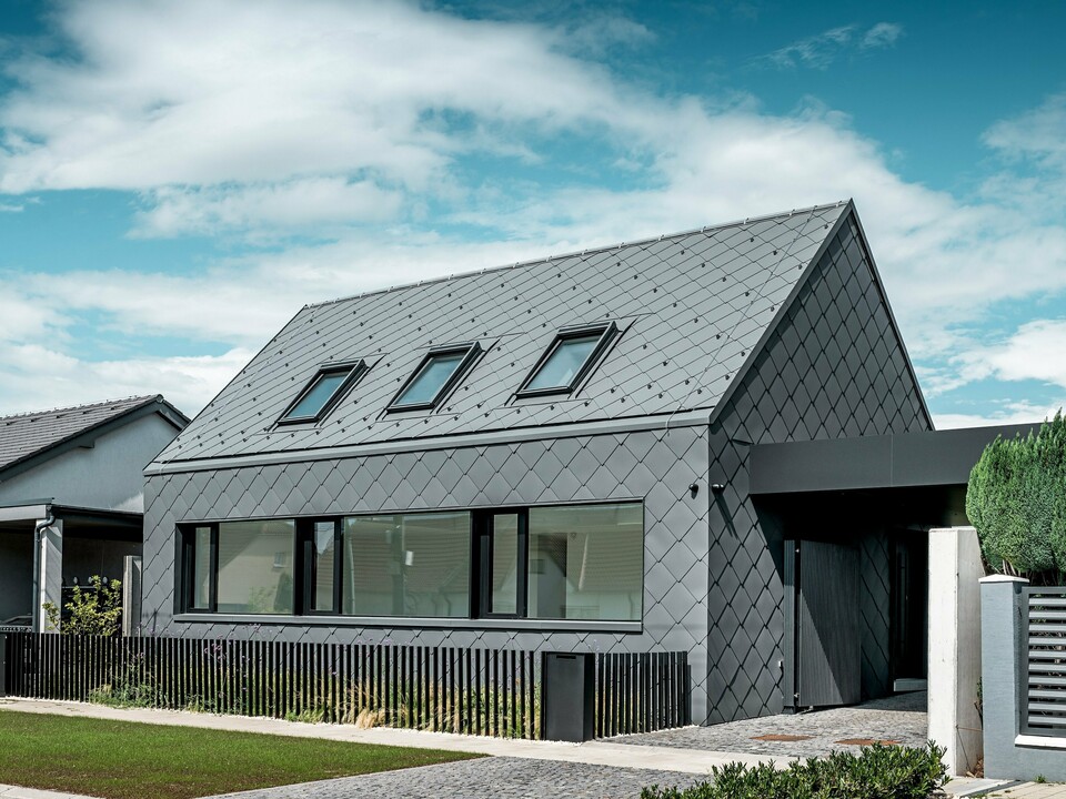 Moderný rodinný dom v Trnave s hliníkovým plášťom: Celý plášť tejto strohej budovy bol vytvorený pomocou strešných a fasádnych šablón PREFA 44 × 44 v odtieni P.10 tmavošedá. Výrazná kosoštvorcová štruktúra plynule prechádza cez strechu a fasádu a dodáva domu silný, monolitický dojem. Veľké okenné otvory vytvárajú jasné architektonické akcenty, zatiaľ čo hliník PREFA odolný voči poveternostným vplyvom ponúka dlhodobú ochranu a ušľachtilý matný vzhľad. Svetlá obloha umocňuje kontrast s tmavým plášťom budovy a zdôrazňuje moderný dizajn.