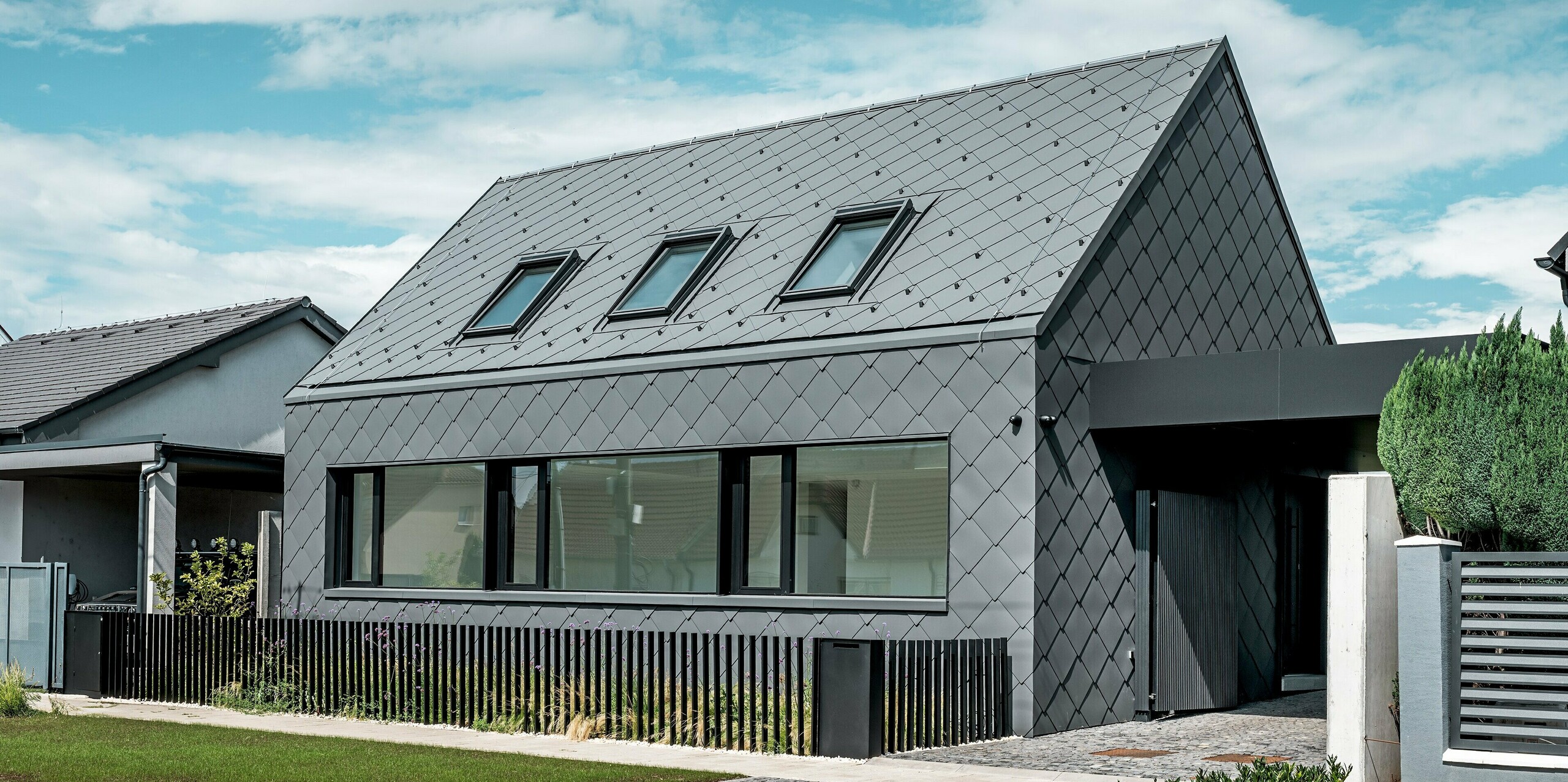 Moderný rodinný dom v Trnave s hliníkovým plášťom: Celý plášť tejto strohej budovy bol vytvorený pomocou strešných a fasádnych šablón PREFA 44 × 44 v odtieni P.10 tmavošedá. Výrazná kosoštvorcová štruktúra plynule prechádza cez strechu a fasádu a dodáva domu silný, monolitický dojem. Veľké okenné otvory vytvárajú jasné architektonické akcenty, zatiaľ čo hliník PREFA odolný voči poveternostným vplyvom ponúka dlhodobú ochranu a ušľachtilý matný vzhľad. Svetlá obloha umocňuje kontrast s tmavým plášťom budovy a zdôrazňuje moderný dizajn.