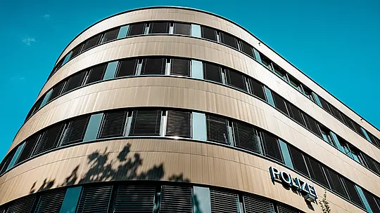 Dramatický pohľad zospodu na zakrivenú novú budovu policajného komisariátu č. 43 v Hamburgu: Výrazná hliníková fasáda PREFA z eloxovaných extrudovaných profilov „Zacke“ v odtieni strednej bronzy sa stáva architektonickým lákadlom. Jemne rýhovaný povrch vytvára živé odlesky svetla a zdôrazňuje plynulé zaoblenia, ako aj horizontálne pásy okien. Vďaka vysokej odolnosti proti korózii, dlhej životnosti a minimálnym nárokom na údržbu ponúkajú hliníkové riešenia PREFA udržateľné, funkčné a esteticky náročné riešenie fasády.