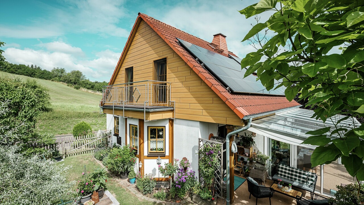 Táto fotografia zobrazuje čarovný rodinný dom v Blieskasteli, ktorého čerstvo zrekonštruovaný štít s PREFA hliníkovými lamelami Siding v odtieni dub prírodny okamžite upúta pozornosť. Teplý vzhľad dreva z odolného hliníka pôsobivo ladí s červenou pálenou strešnou krytinou a priestrannou zimnou záhradou.  Obklopené bujnou zeleňou a starostlivo upravenými kvetinovými záhonmi vzniká idylická, živá scéna. Robustná renovácia fasády s produktmi PREFA dodáva domu nielen prirodzený vzhľad, ale presviedča aj odolnosťou voči poveternostným vplyvom, stálosťou farieb a takmer bezúdržbovou životnosťou.   