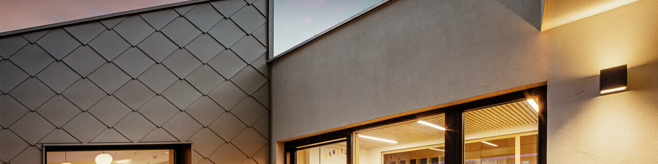 Večerný pohľad na časť školiaceho strediska v obci Gerlos s umeleckou fasádou. Vľavo je okno obložené PREFA fasádnymi šablónami 44x44, vpravo je vchod s veľkorysým okenným priečelím vedúcim do dobre osvetleného interiéru so stoličkami a stolmi. Teplé osvetlenie interiéru kontrastuje s matným svetlom súmraku a zvýrazňuje modernú architektúru a dizajn fasády.