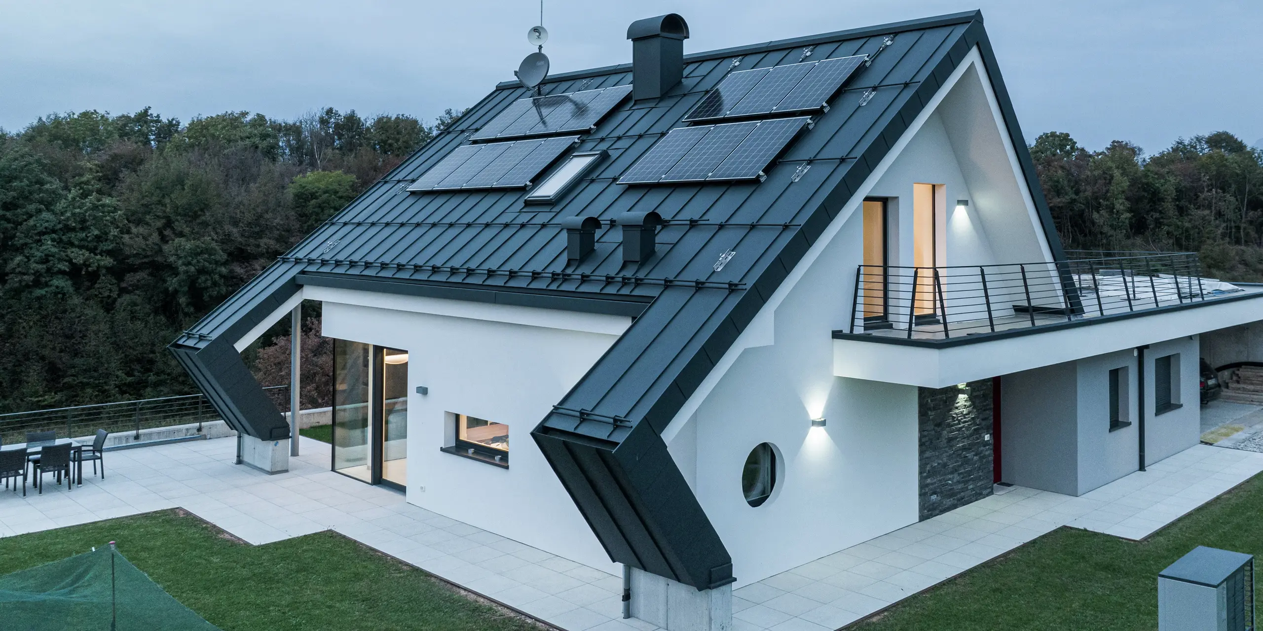Moderný rodinný dom v Borgo Valbelluna so strešnou krytinou PREFALZ vo farbe P.10 antracit. Hliníková strecha s fotovoltickými modulmi spája udržateľnú výrobu energie s nadčasovou estetikou. Nápadné presahy strechy zdôrazňujú inovatívny dizajn a poskytujú dodatočnú ochranu pred poveternostnými vplyvmi. Veľké sklenené plochy, bielo omietnutá fasáda a balónový záves vytvárajú harmonický celkový vzhľad.