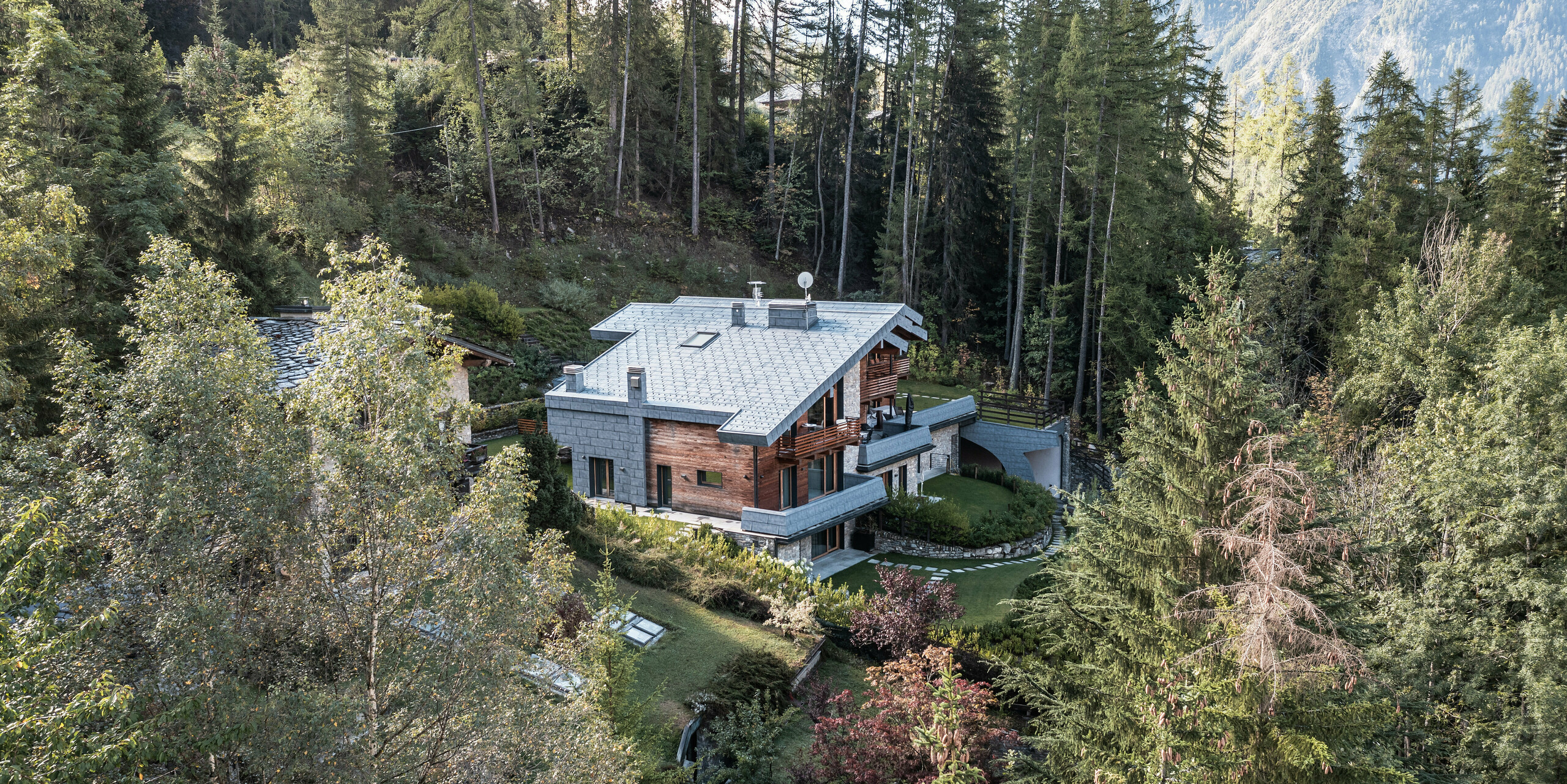 Letecký pohľad na impozantnú vilu v Courmayeur, obklopenú hustými lesmi zvýrazňujúci prírodnú architektúru z dreva, kameňa a PREFA hliníka. Strešné a fasádne panely FX.12 vo farbe kamenná šedá od PREFA pôsobia ako pútače a poskytujú budovu dlhodobú ochranu. Stupňovité usporiadanie stavebných častí umožňuje integráciu do strmej krajiny, zatiaľ čo niekoľko balkónov a dobre udržiavaných záhrad ponúka príjemnú vonkajšiu plochu, ktorá pozýva na odpočinok v pokojnom alpskom prostredí.