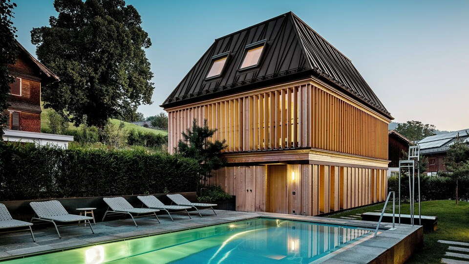 „Kúpeľný dom“ hotela Hirschen v Schwarzenbergu za súmraku, s výhľadom na osvetlený vonkajší bazén v popredí. Budova zaujme svojou modernou valbovou strechou z PREFALZ vo farbe P.10 orieškovohnedá s presne vyhotovenými, čiastočne šikmými stojatými falcami. Dve strešné okná zabezpečujú denné svetlo vo vnútri. Fasáda sa skladá z vertikálnych drevených lamiel po celej výške poschodia, ktoré dodávajú budove štruktúru a ľahkosť. V spodnej časti dominuje prírodné drevo. Obklopený upravenou zeleňou a zasadený do kopcovitej krajiny Bregenzerwaldu vytvára harmonické spojenie architektúry, prírody a oddychu.