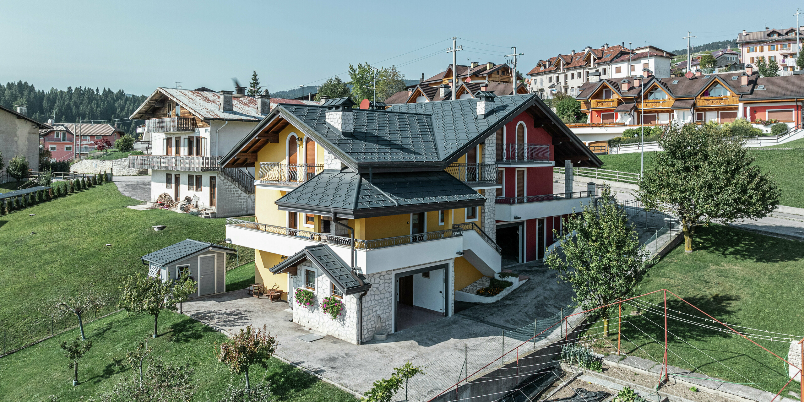 Rodinný dom v Gallio v Taliansku v tradičnom alpskom štýle s modernou strechou PREFA v tmavošedej farbe P.10. Strešné panely PREFA R.16 poskytujú optimálnu ochranu pred snehom, dažďom a vetrom. Jedinečná kombinácia žlto-červenej fasády, prvkov z prírodného kameňa a drevených akcentov dodáva domu príjemný vzhľad. Strešný odvodňovací systém PREFA zaisťuje spoľahlivé odvádzanie zrážok. Tento dom, zasadený do zeleného kopcovitého krajiny, spája alpský šarm s odolnou strešnou technikou, ktorá nevyžaduje veľa údržby.