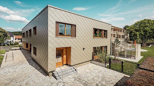 Obrázok zobrazuje materskú školu v Alzenau zo šikmej vtáčej perspektívy a zdôrazňuje modernú architektúru s výraznou bronzovou hliníkovou fasádou PREFA. Fasáda je tvorená fasádnymi šablónami 44 × 44, ktoré vytvárajú harmonický šupinový vzor a dodávajú stavebnému objemu elegantnú dynamiku. Veľké drevené okná s vertikálnymi lamelami prinášajú teplé akcenty a prepúšťajú do interiéru množstvo denného svetla. Na pravej strane obrázka sa otvára areál s oploteným ihriskom, zelenými plochami a krytou terasou. Kombinácia trvácneho, bezúdržbového hliníka a udržateľných materiálov robí z budovy vzorový projekt funkčnej a ekologickej architektúry v oblasti vzdelávacích stavieb.