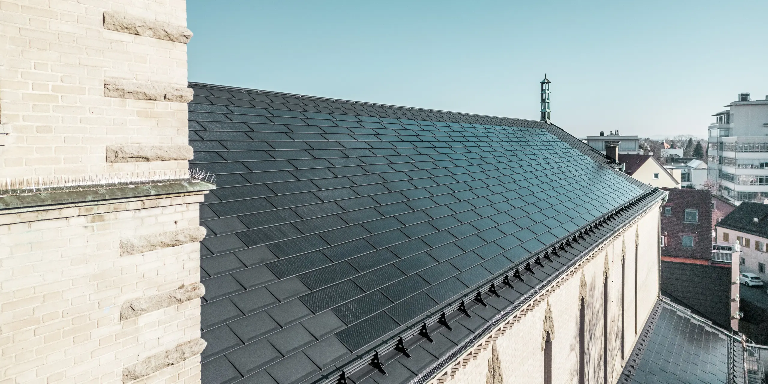 Pohľad za kostolnú vežu ukazuje integrované solárne panely PREFA v elegantnej čiernej farbe na streche kostola svätého Karola Boromejského v Norimbergu. Perspektíva zdôrazňuje dokonalú integráciu solárnych modulov, ktoré bez problémov zapadajú do sedlovej strechy pamiatkovo chránenej budovy. Rozpoznateľné sú aj výrazné dekoratívne prvky na stene kostola, ktoré zdôrazňujú franskú expresionistickú architektúru. Integrované solárne panely zabezpečujú udržateľnú energiu pre kostol a susedné budovy a zároveň predstavujú estetické riešenie v oblasti pamiatkovej ochrany.
