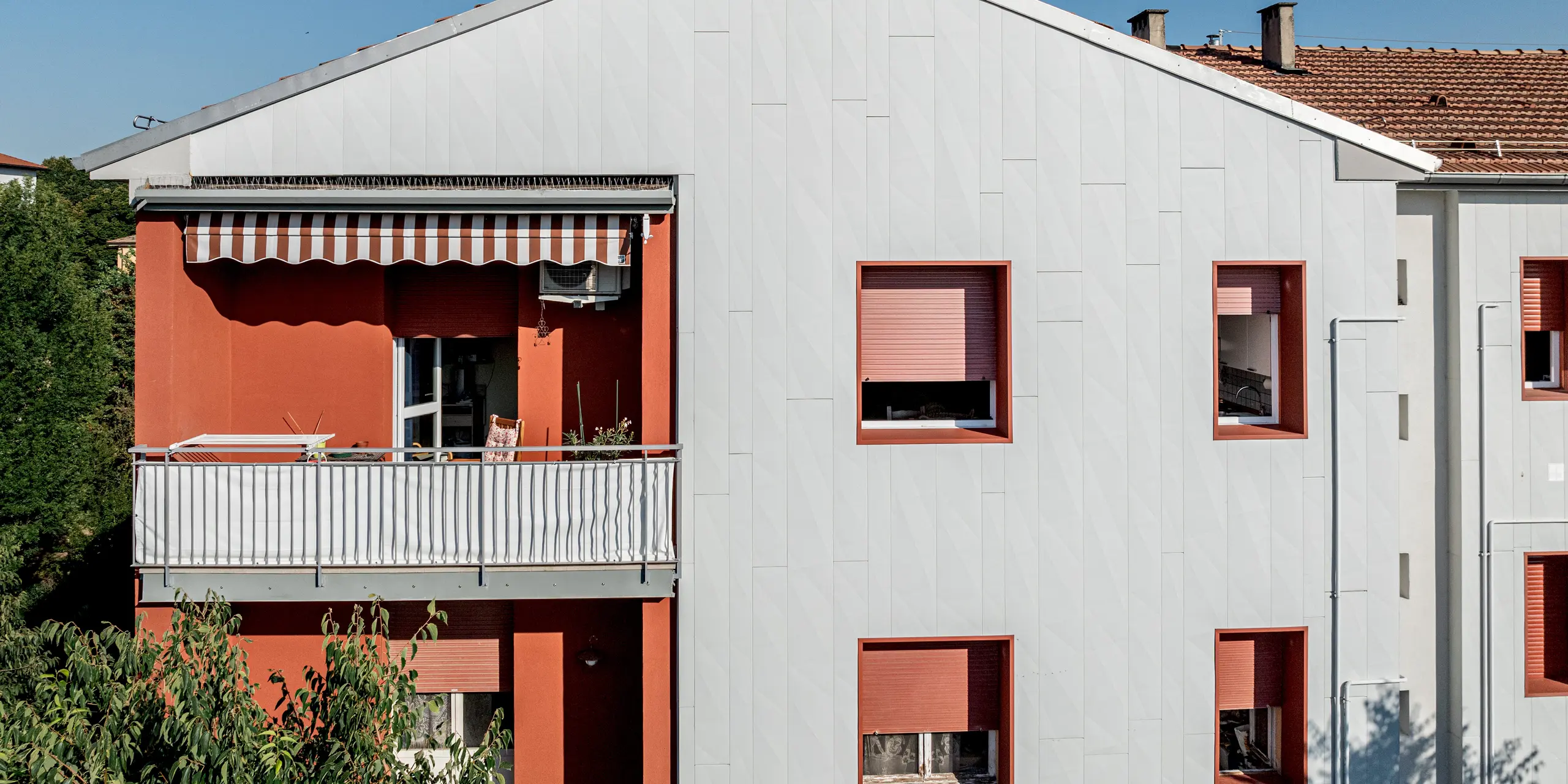 Detailansicht der sanierten Wohnanlage Alba B mit markantem Giebel und klar gegliederter Fassadenfläche aus PREFA Siding.X Aluminiumpaneelen. Die vertikal verlegten Paneele erzeugen eine feine, lebendige Struktur mit dezenten Licht- und Schattenspielen. Links kontrastiert ein Balkon mit rot verputzter Wand und Markise, rechts setzen tief eingeschnittene Fenster mit roten Laibungen starke Akzente. Die PREFA Aluminiumfassade überzeugt durch hohe Witterungsbeständigkeit, Langlebigkeit und wartungsarme Qualität bei moderner Optik.