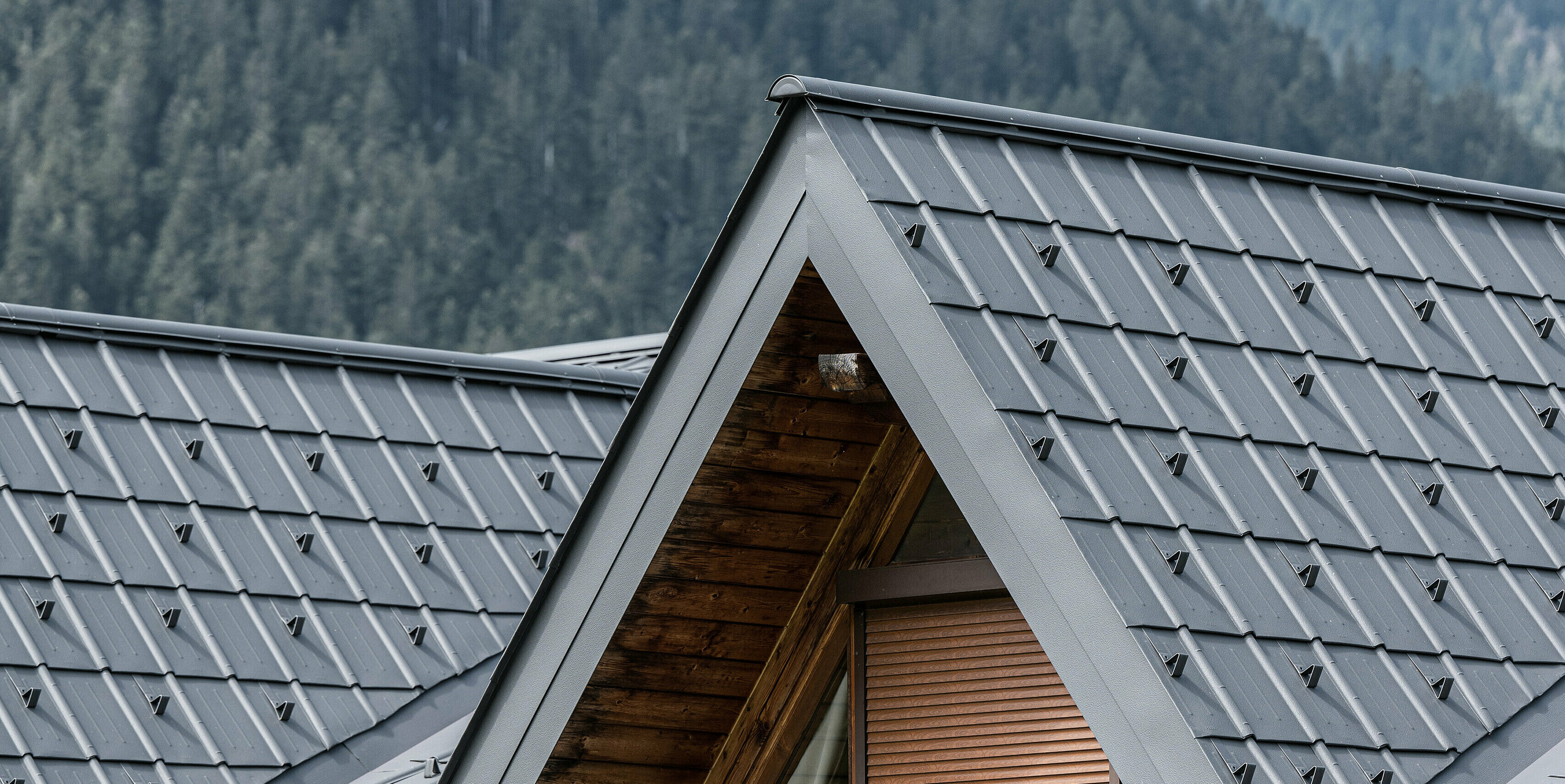 Detail vikiera na alpskom dome v Auronzo di Cadore, ktorý je vybavený škridlami PREFA v svetlošedej farbe. Jemne štruktúrované hliníkové škridle nádherne kontrastujú s prírodným drevom strešnej konštrukcie, poskytujú vysokokvalitnú ochranu pred alpským počasím a zároveň dokonale zapadajú do horskej lesnej krajiny.