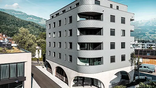 Vonkajší pohľad na Bernerhaus v Buchsi s výrazným trojuholníkovým tvarom a jemne zaoblenými balkónovými pásmi. Fasáda z PREFA fasádnych šindľov v odtieni P.10 svetlošedá obklopuje budovu ako šupinatá hliníková koža a vytvára presný, živý obraz fasády. Nad masívnym betónovým podstavcom s elegantnými arkádami pôsobí ľahká, odolná proti poveternostným vplyvom hliníková fasáda PREFA obzvlášť výrazne. Trvanlivá, nenáročná na údržbu a tvarovo stabilná, vytvára moderný architektonický dojem na pozadí alpských vrcholov.