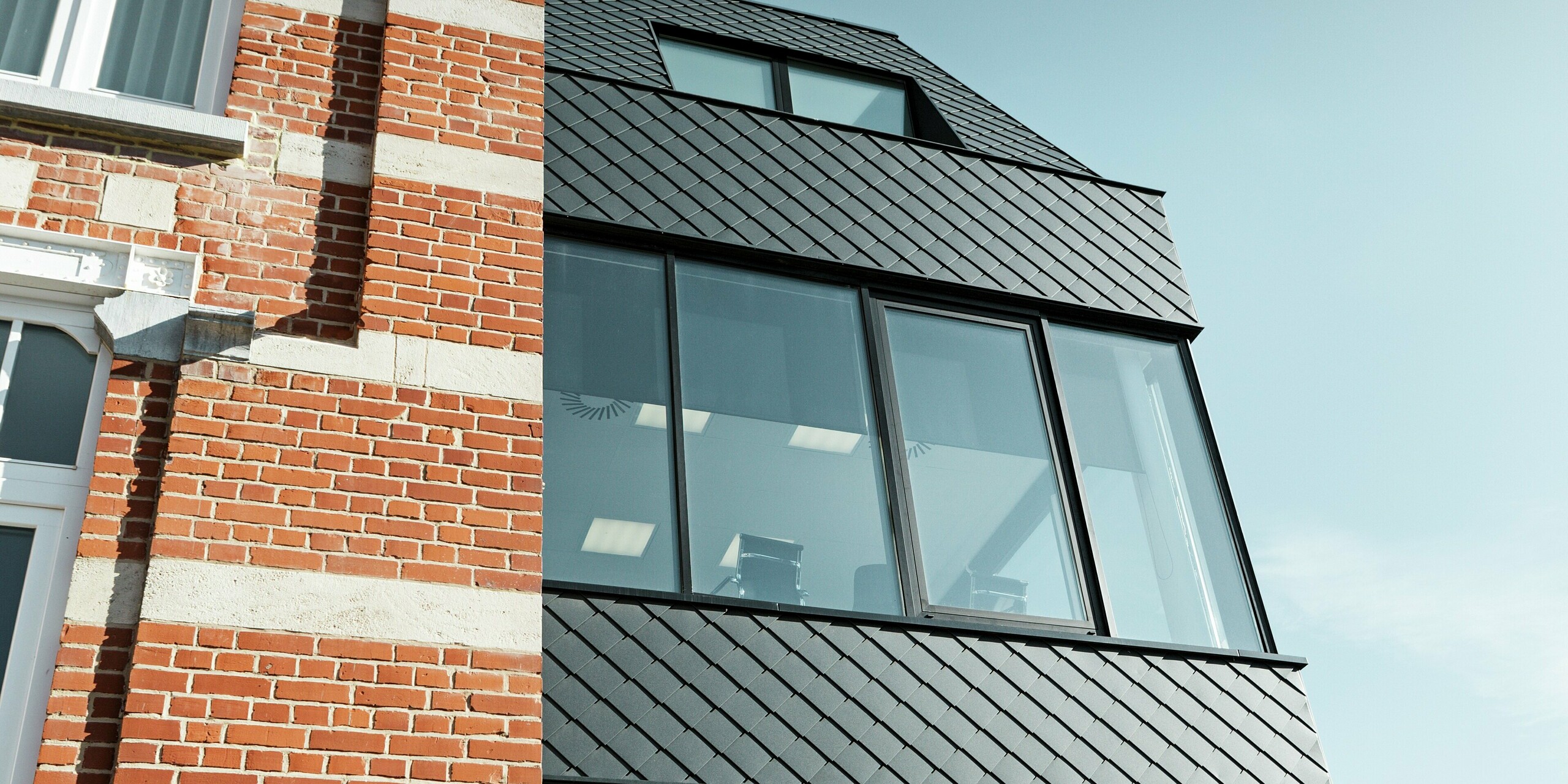 Nahaufnahme des Kasteeltje Heizel: Der rote Backstein mit Natursteinfundament trifft direkt auf den modernen Zubau mit anthrazitfarbenen PREFA Wandrauten und großen Glasflächen. Die diagonale Linienführung der Aluminiumfassade setzt einen starken Kontrast zum historischen Mauerwerk und macht die Verbindung von Tradition und zeitgemäßer Architektur eindrucksvoll sichtbar.