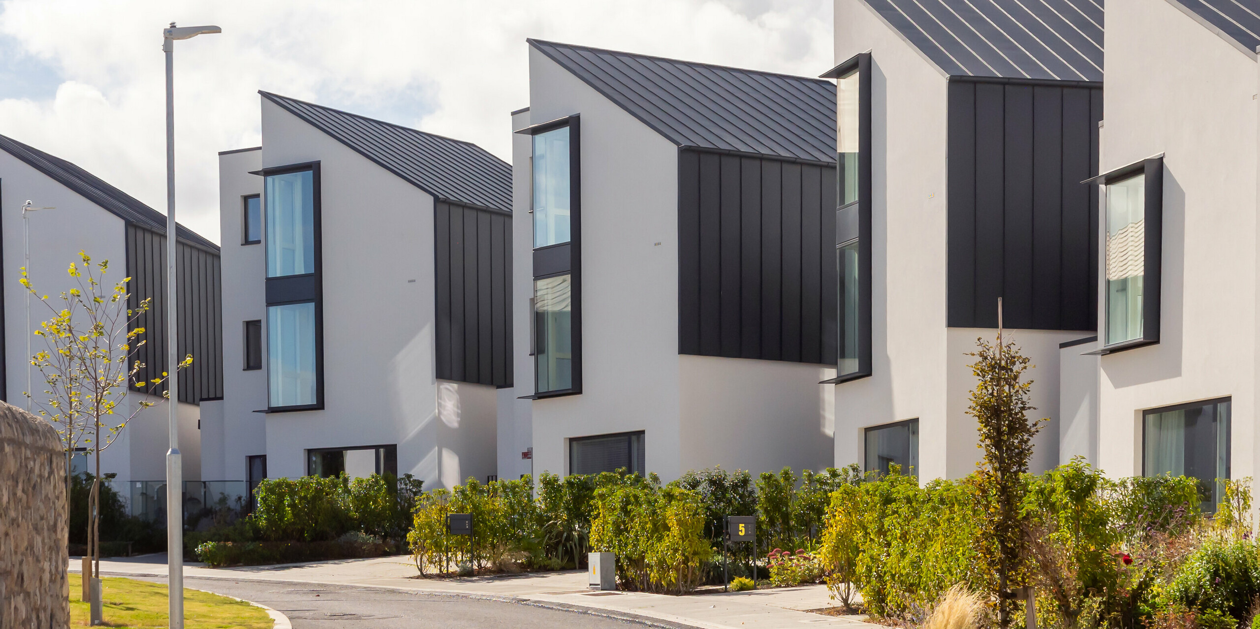 Moderne Reihenhausanlage Watson Place in der Nähe von Dublin mit klaren, asymmetrischen Dachformen und dunkler PREFALZ Verkleidung in P.10 Anthrazit. Die großformatigen Fenster und die markanten Aluminiumflächen betonen die dynamische Architektur. Die langlebigen PREFA Produkte bieten zuverlässigen Witterungsschutz und verleihen der Wohnanlage eine elegante, hochwertige Erscheinung.