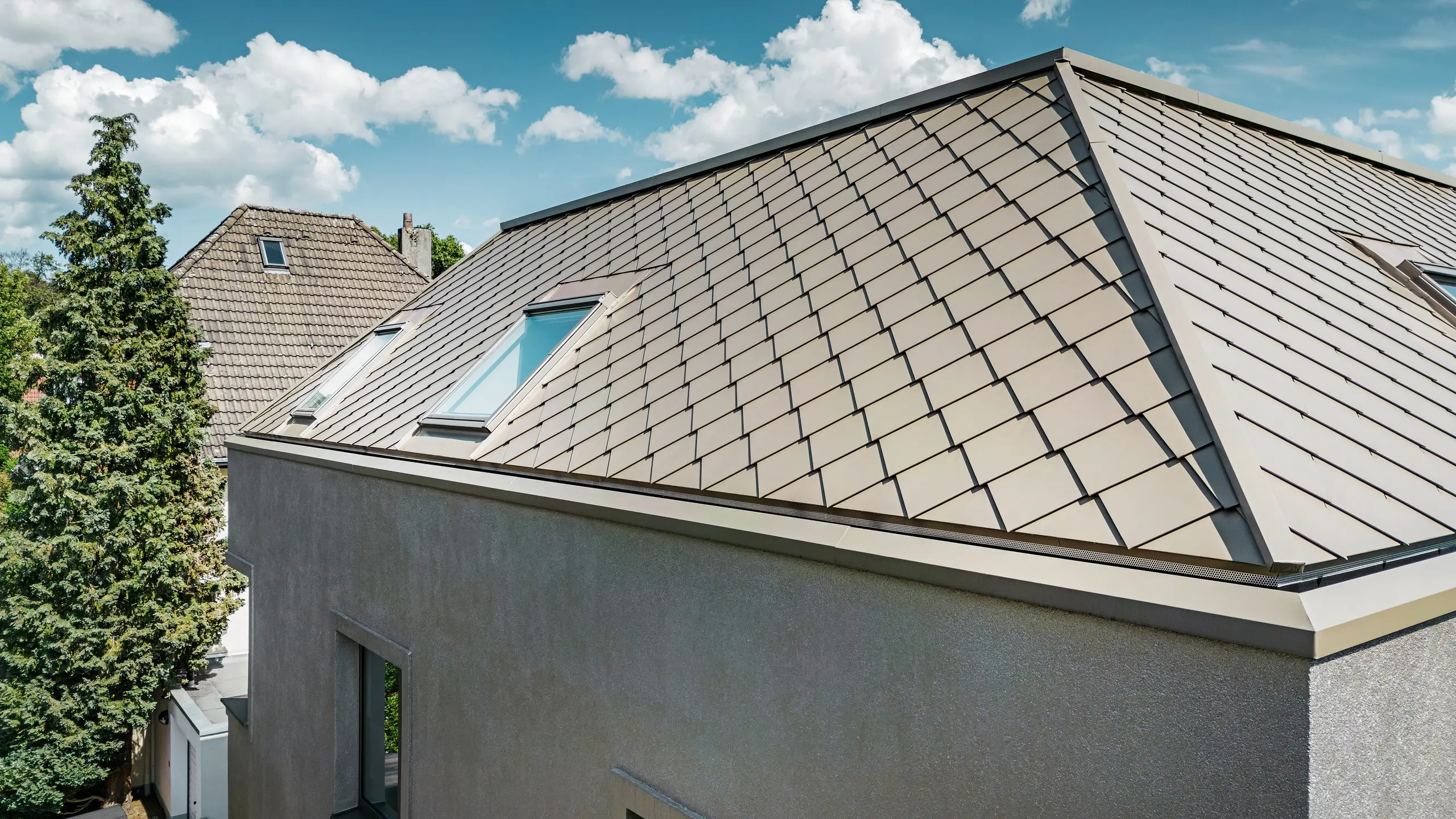 Detailný záber na valbovú strechu moderného rodinného domu s hliníkovým strešným plášťom bronzovej farby od spoločnosti PREFA. Strešná konštrukcia vykazuje vysokokvalitnú povrchovú úpravu s integrovanými strešnými oknami, ktoré poskytujú dodatočné prirodzené svetlo v interiéri. Čisté línie a lesklý povrch hliníkových strešných panelov zdôrazňujú moderný a elegantný dizajn domu.