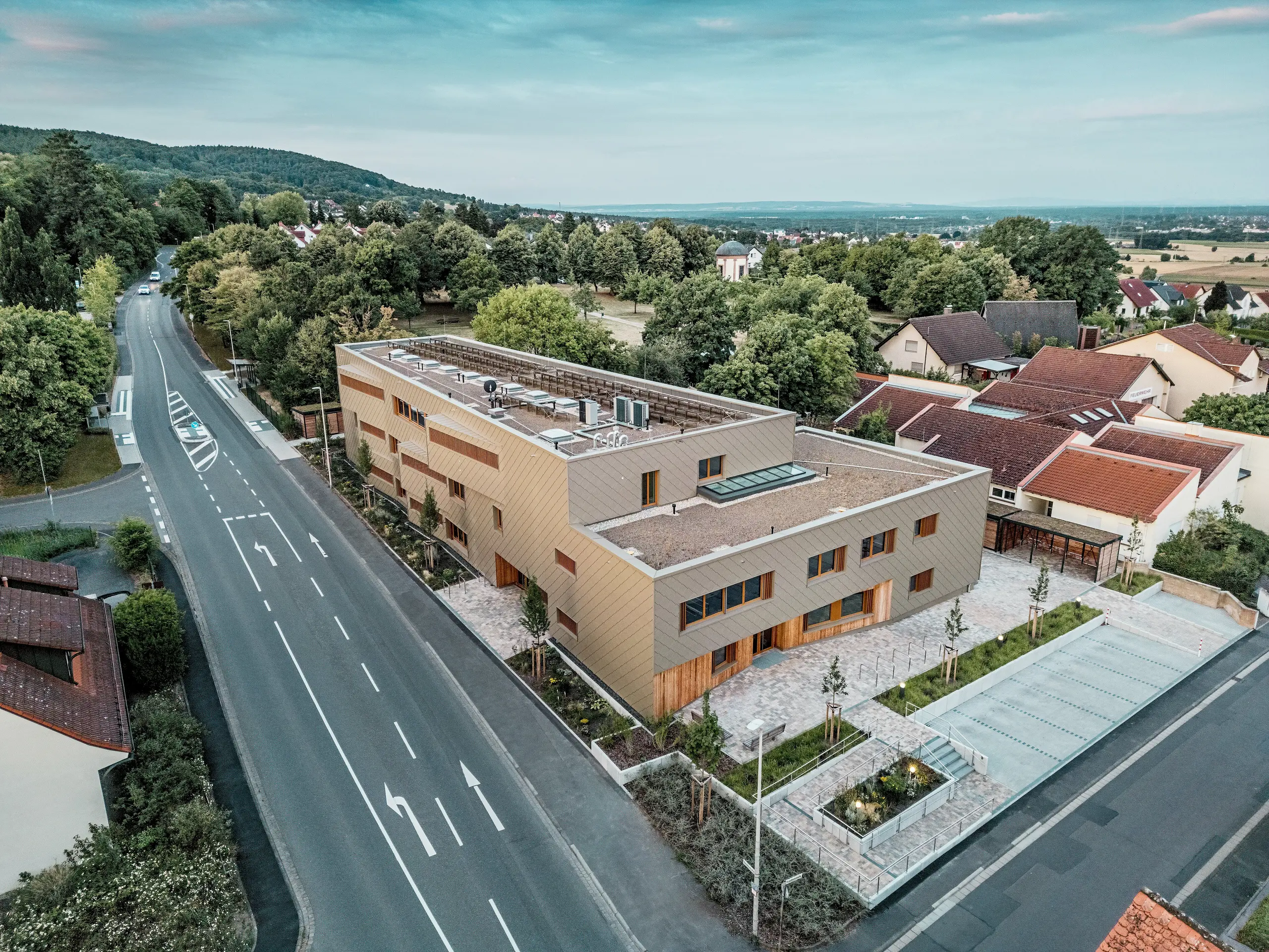 Impozantné celkové riešenie materskej školy v Alzenau je viditeľné z leteckého pohľadu. L tvarová hybridná budova je harmonicky zasadená do pokojnej obytného štvrti na okraji Spessartu. Fasáda z bronzovo sfarbených PREFA fasádnych šablón 44 × 44 dodáva stavbe výrazný a kvalitný vzhľad a zdôrazňuje modernú architektúru. Zelené strechy, priestranné vonkajšie plochy a jasné riešenie dopravy podčiarkujú udržateľný koncept celku. Kombinácia trvácneho hliníka, dreva a recyklovaného betónu spĺňa najvyššie ekologické štandardy. Táto perspektíva zvýrazňuje premyslenú urbanistickú integráciu a multifunkčný charakter budovy – perspektívny projekt pre vzdelávanie, bývanie a udržateľnosť.