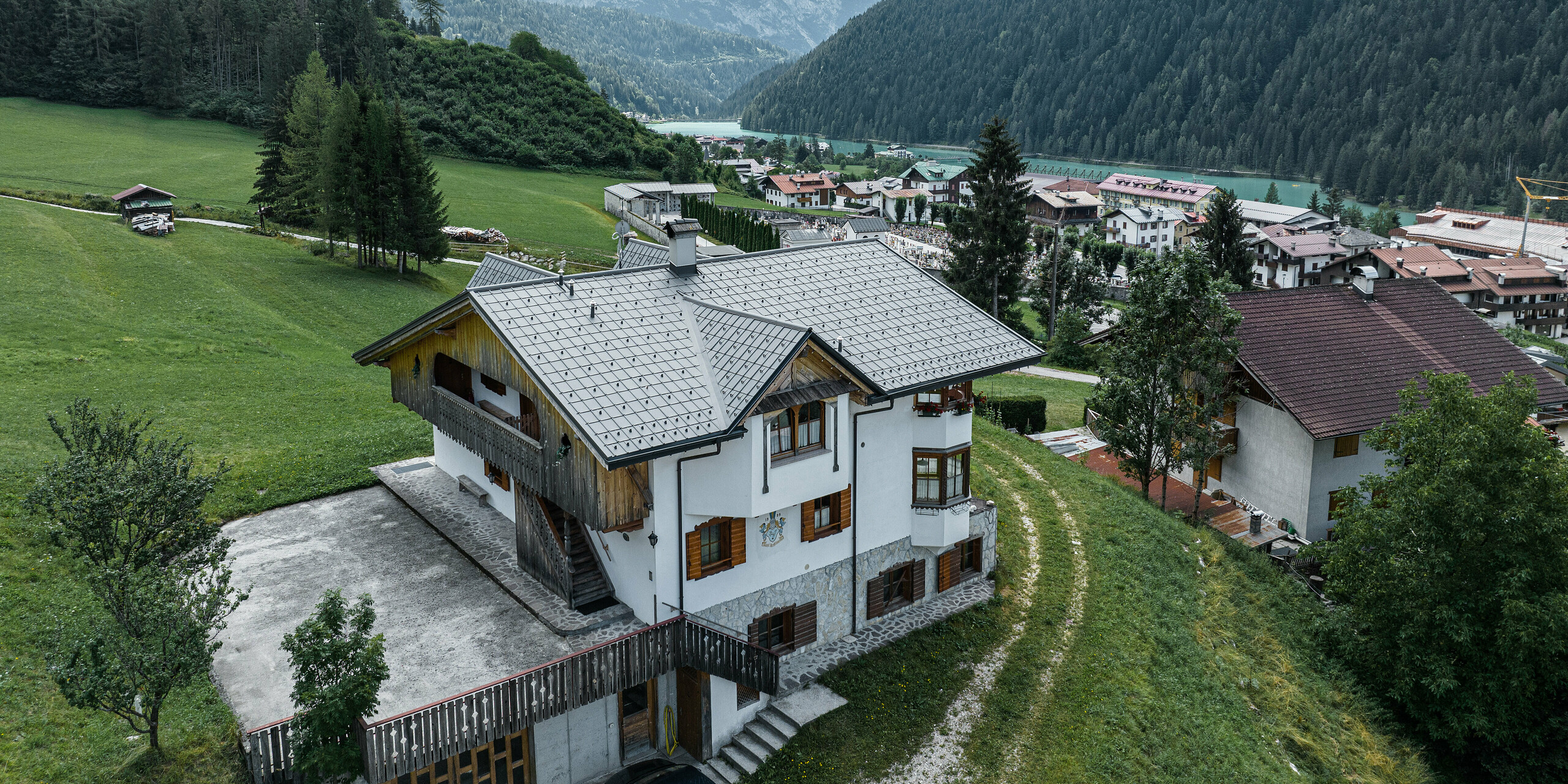 Letecký pohľad na pôvabnú horskú chatu v Auronzo di Cadore v Taliansku po renovácii strechy s hliníkovými škridlami PREFA v svetlosivej farbe. Chata sa týči na zelenej lúke s pôsobivým horským masívom v pozadí a ukazuje malebnú súhru modernizovaných stavebných prvkov a tradičnej alpskej architektúry. Nad vstupným priestorom je vidieť špicatý vikier s dreveným štítom.