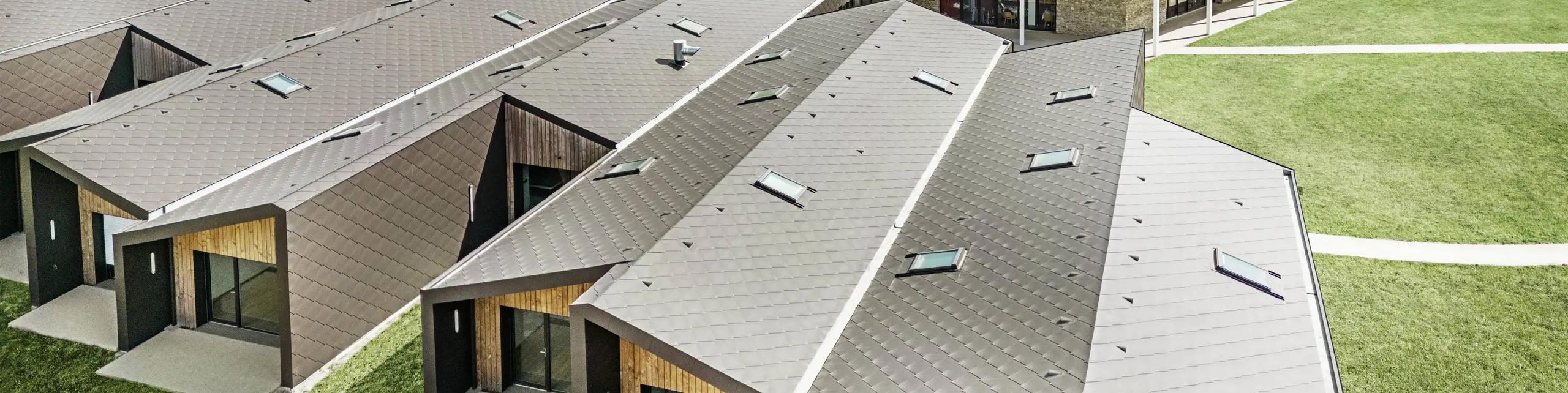 Na obrázku je letecký pohľad na komplex moderných budov obložených strešnými a fasádnymi šablónami PREFA 44 × 44 vo farbe orieškovohnedá. Strešnú krajinu charakterizujú geometrické tvary v kombinácii s veľkými sklenenými priečeliami a drevenými prvkami. V pozadí je vidieť starý kostol, ktorý tvorí harmonický kontrast k modernej architektúre. Zelený trávnik a okolitá príroda zdôrazňujú otvorený a udržateľný dizajn celého komplexu.