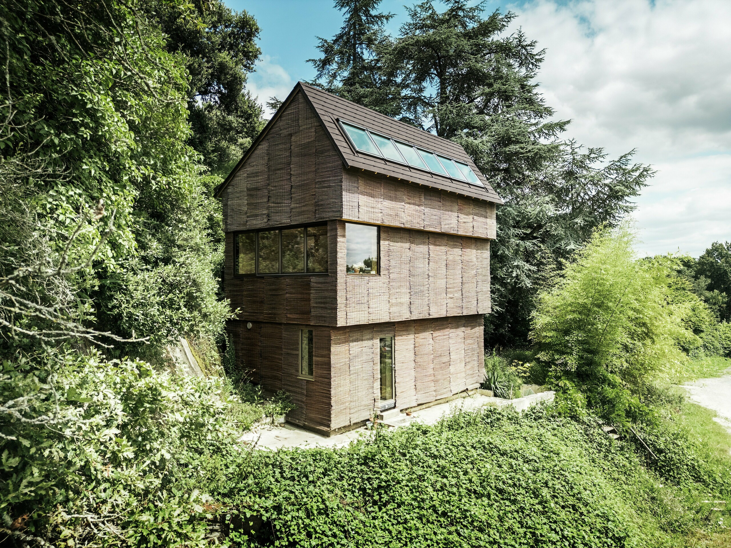 Dom vo Francúzsku so strešnými šindľami PREFA v orieškovohnedej farbe P.10 a vŕbovou fasádou, obklopený prírodnou zeleňou - udržateľná architektúra a ekologický dizajn.
