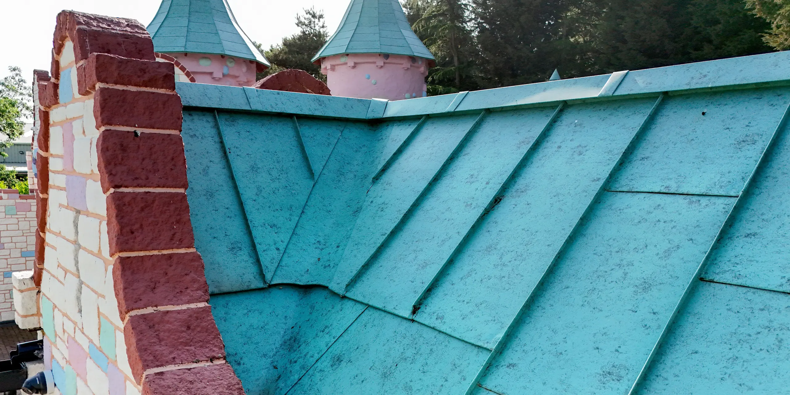 Detailaufnahme eines kunstvoll ausgeführten PREFALZ Stehfalzdachs in P.10 Patinagrün im Sundown Adventureland, das mit präzise gearbeiteten Anschlüssen und sauber ausgeführten Übergängen überzeugt. Die markanten Falzlinien betonen die dynamische Dachform und setzen einen starken visuellen Akzent neben den farbigen Ziergiebeln. Besonders bei komplexen Geometrien zeigt das PREFA Aluminium seine hohe Formbarkeit und ermöglicht exakte Lösungen. Gleichzeitig punktet das Material durch Korrosionsbeständigkeit, Langlebigkeit und zuverlässigen Schutz bei jedem Wetter.
