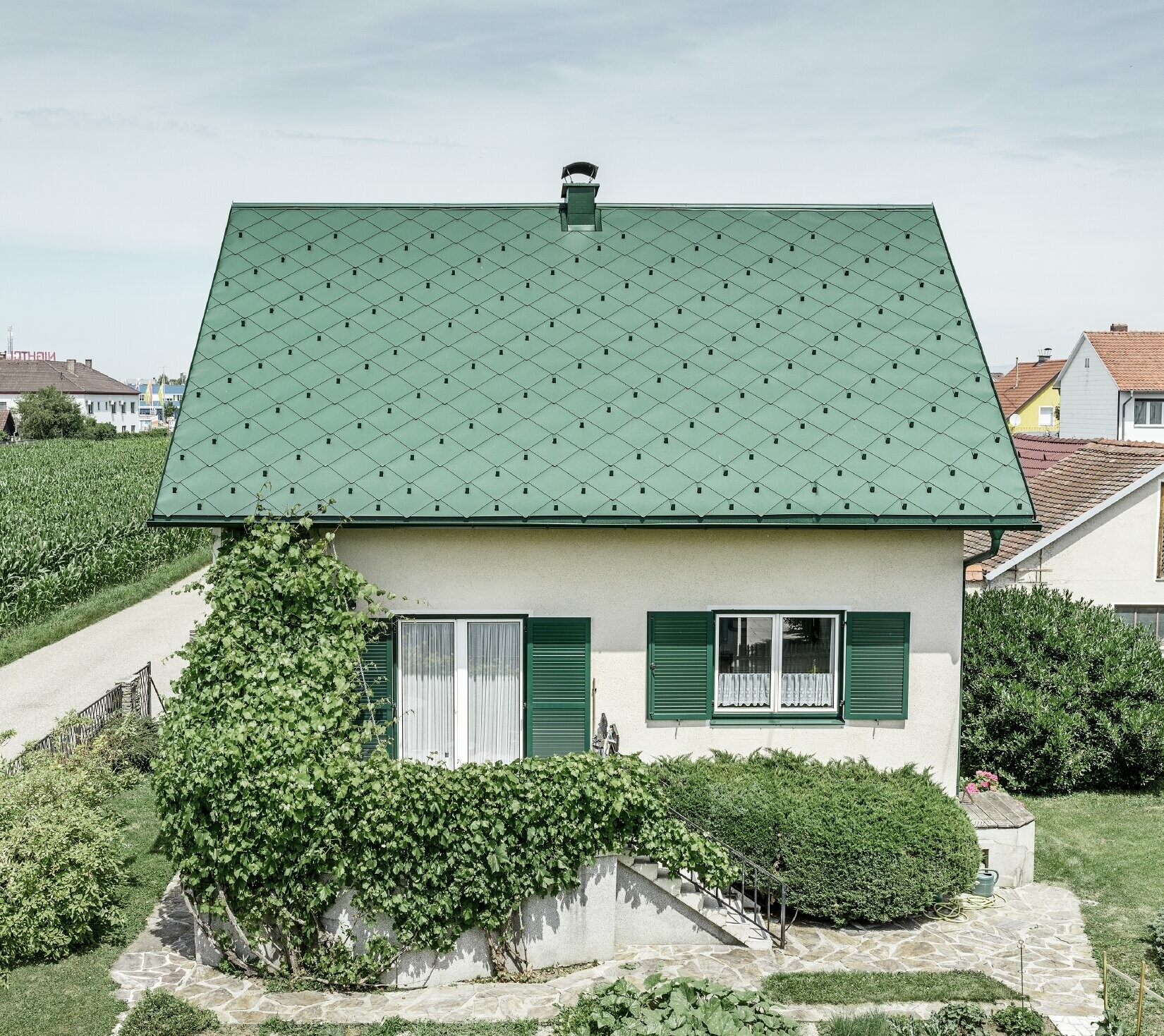 Klasický rodinný dom so sedlovou strechou so strešnou krytinou z hliníka vo farbe machovozelená so zelenými okenicami. Strecha bola pokrytá PREFA strešnými šablónami 44 × 44 vo farbe P.10 machovozelená.