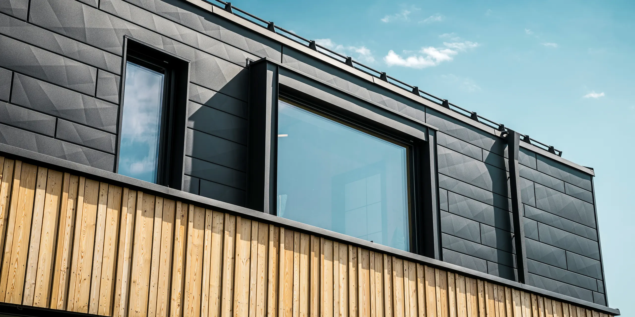 Detailný pohľad na horné poschodie vzorového domu Autark v centre prefabrikovaných domov Blaue Lagune, Vösendorf (AT). Fasáda je tvorená hliníkovými panelmi PREFA Siding.X v čiernej farbe P.10, ktoré zaujímavým spôsobom vynikajú svojím špeciálnym vzhľadom s rôznymi ohybmi. Pod nimi sa nachádza štýlové drevené obloženie, ktoré vytvára harmonický kontrast s tmavým hliníkom. Veľké okná prepúšťajú do interiéru veľa svetla a zdôrazňujú moderný dizajn.