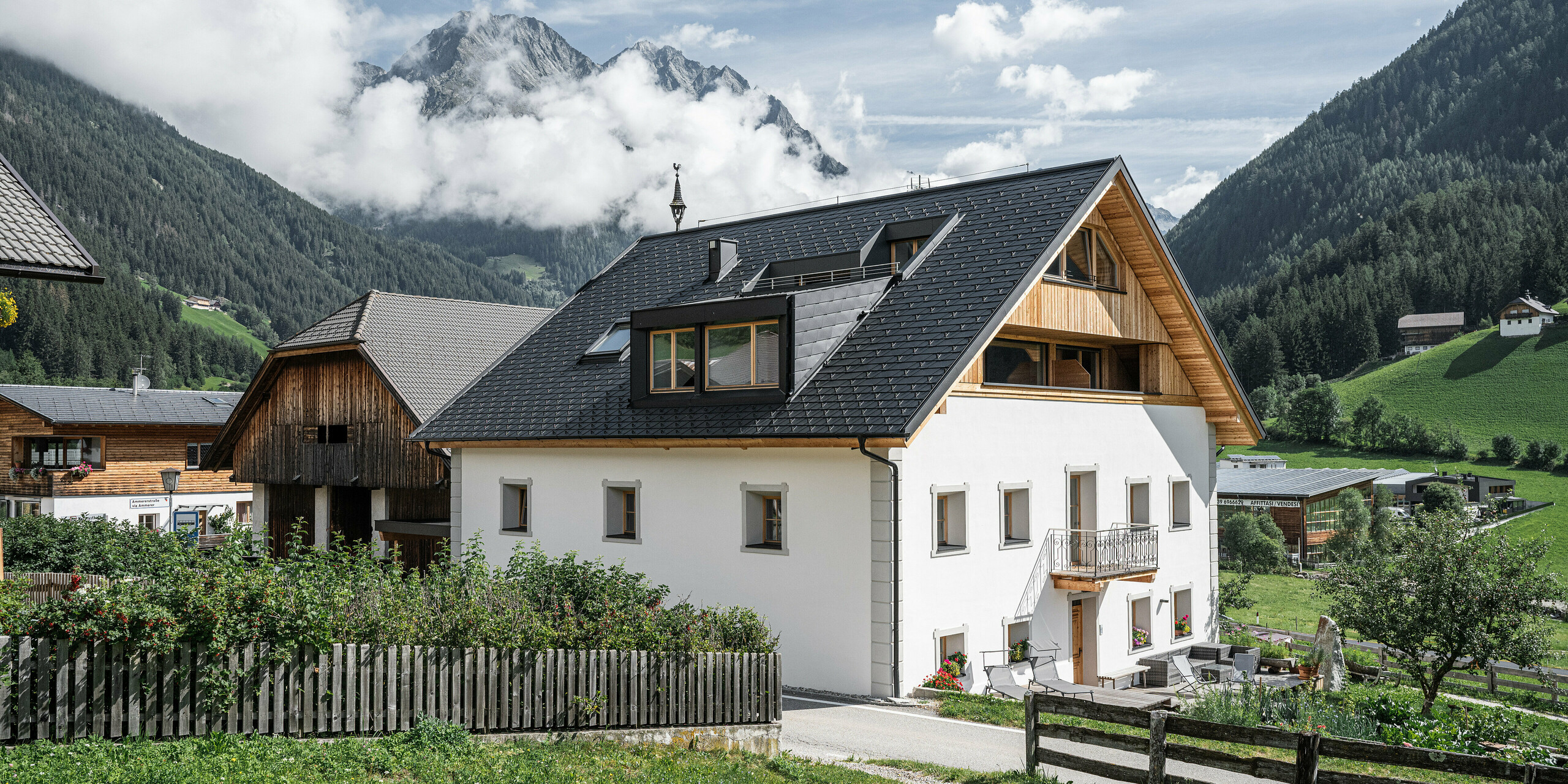 Celkový pohľad na rekreačné apartmány Ausserwegerhof v idylickom údolí Antholz, Južné Tirolsko, pokryté strešným šindľom PREFA DS.19 v antracitovej farbe. Tradičná stavba sa snúbi s modernými prvkami a ponúka malebný výhľad na majestátnu horskú kulisu, tajomne obklopenú mrakmi. Ausserwegerhof v strede obrázku je obklopený klasickými farmami a zelenými lúkami.