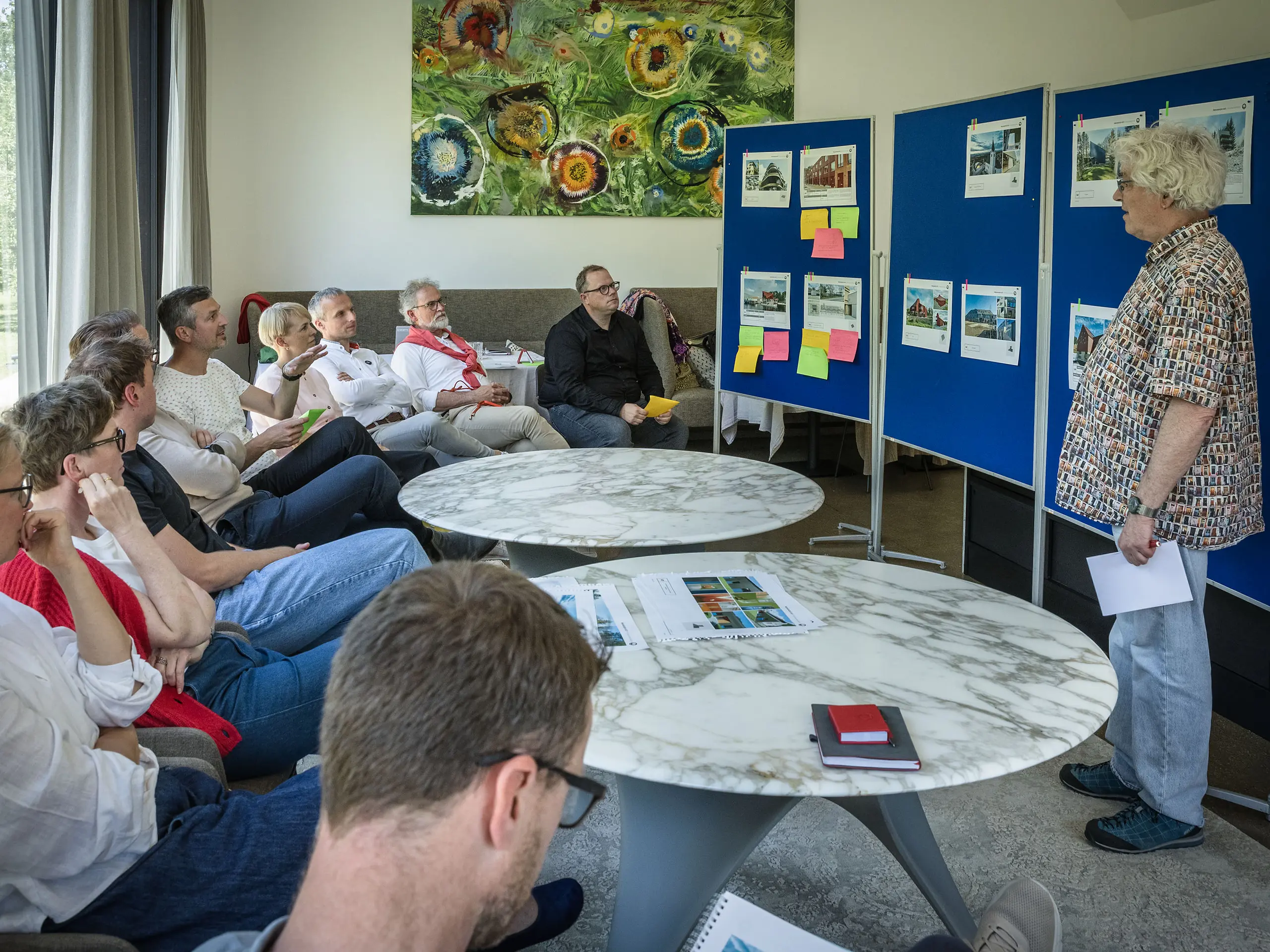 PREFARENZEN 2027 Zasadnutie poroty v svetlej seminárnej miestnosti: Moderátor Matthias Finkentey prezentuje pred modrými nástenkami architektonické projekty z hliníka PREFA, zatiaľ čo porota pozorne sedí a robí si poznámky. Veľké okná vpúšťajú dovnútra denné svetlo a ponúkajú výhľad na zelenú záhradu. Na stoloch ležia knihy o architektúre, zošity a perá. Scéna vyžaruje sústredenú analýzu, kreatívnu výmenu názorov a hodnotenie dizajnu a funkčnosti. Hliníkové produkty PREFA symbolizujú trvácne, inovatívne strechy a fasády, ktoré spájajú estetiku, kvalitu a inšpiráciu pre architektonické projekty.