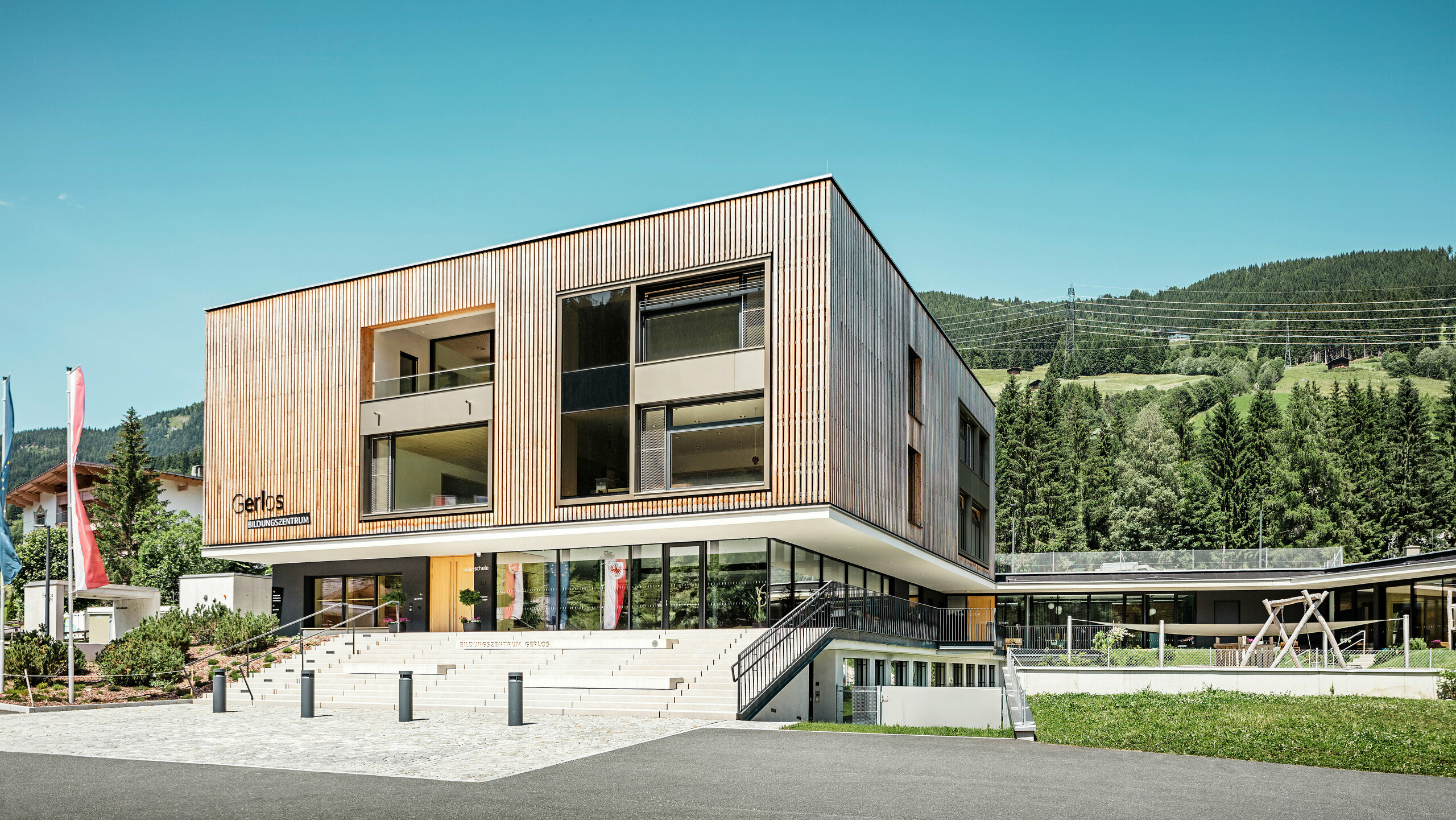 Pohľad na moderné vzdelávacie centrum s drevenou fasádou spredu. Budova je dvojpodlažná s veľkými okennými priečeliami a plochou strechou, ktorá dokonale zapadá do okolitej horskej krajiny husto zalesnených kopcov. V popredí sú upravené zelené plochy, stupňovitá príjazdová cesta a reklamné vlajky. Návrh vyjadruje kombináciu funkčnosti a estetickej architektúry v harmonickom súlade s prírodou.