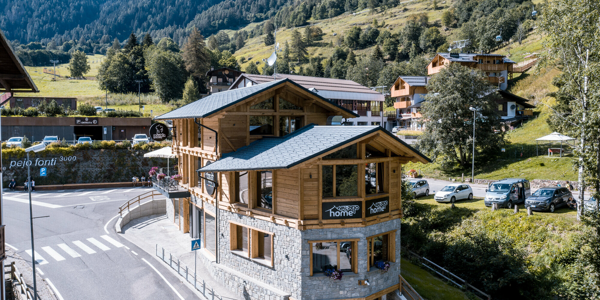 Pohľad na moderný après-ski bar v tradičnej drevenej architektúre s kamenno-sivými strešnými šindľami PREFA DS.19, vyrobenými z hliníka, začlenený do idylickej horskej krajiny so sviežou zeleňou a výhľadom na Alpy.