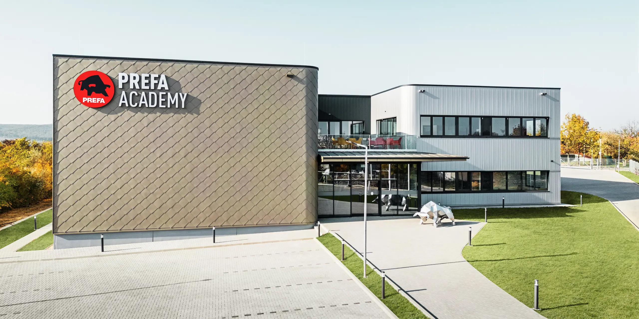 Nové sídlo spoločnosti PREFA Hungary v Törökbálinte zaujme modernou fasádou z fasádnych šablón PREFA 44 × 44 v bronzovej farbe a hliníkových lamiel PREFA Siding vo farbe strieborná metalíza. Odolné hliníkové výrobky zabezpečujú odolnosť voči poveternostným vplyvom, nenáročnú údržbu a vizuálne príťažlivý plášť budovy. Výrazné logo PREFA a plastika hliníkového býka zdôrazňujú inovatívnu silu, kvalitu a pevnosť spoločnosti.