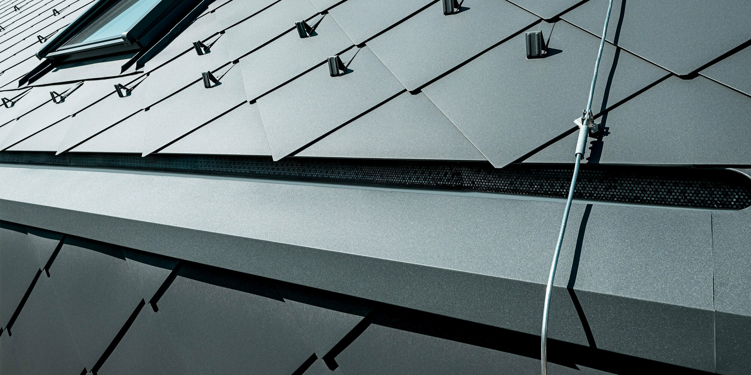 Detailný záber strechy PREFA so strešnými šablónami 44 × 44 v odtieni P.10 tmavošedá na rodinnom dome v Trnave. Na snímke je vidieť precízne položenú hliníkovú krytinu s funkčnými snehovými zábranami a integrovaným strešným oknom. Matný povrch šablón rovnomerne odráža svetlo a zdôrazňuje vysokú kvalitu spracovania. Trvanlivé hliníkové produkty PREFA ponúkajú maximálnu stabilitu, odolnosť voči poveternostným vplyvom a moderný, technicky pôsobiaci vzhľad, ktorý budove dodáva jej výraznú architektúru.