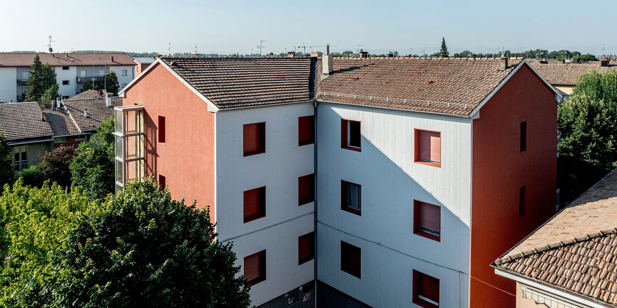 Blick über die sanierte Wohnanlage Alba B mit klar gegliederten Baukörpern und Satteldächern, eingebettet in eine gewachsene Wohnumgebung mit dichter Begrünung. Die Fassade aus PREFA Siding.X Aluminiumfassadenpaneelen hebt sich durch ihre helle, strukturierte Oberfläche deutlich ab, während kräftig rote Fensterlaibungen markante Akzente setzen. Einfallendes Licht erzeugt feine Schattenspiele auf den Paneelen und betont die vertikale Verlegung. Die PREFA Aluminiumfassade überzeugt durch Langlebigkeit, Witterungsbeständigkeit und eine dauerhaft wartungsarme Gebäudehülle.