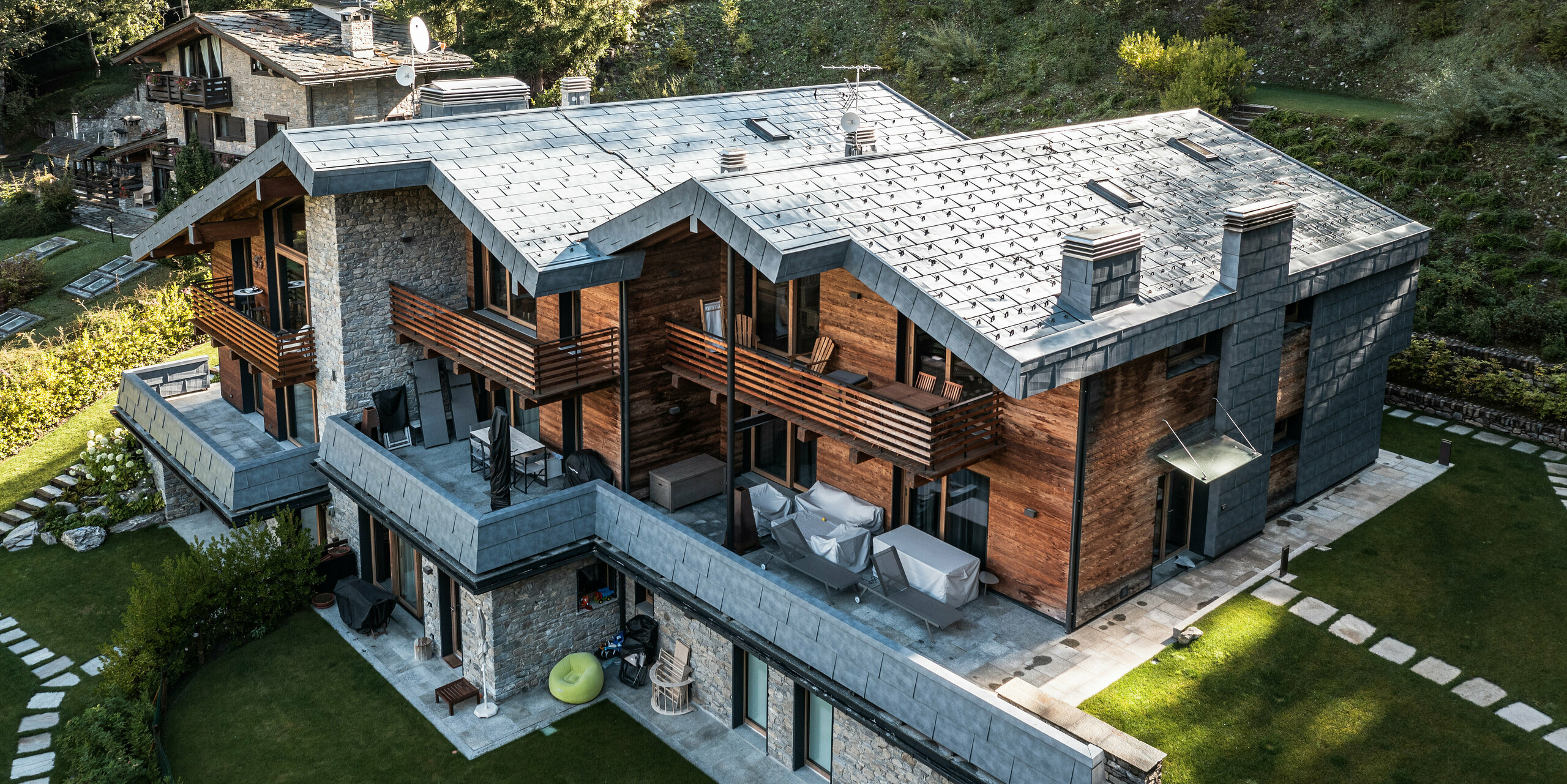 Letecký pohľad na impozantnú vilu v Courmayeur, obklopenú hustými lesmi zvýrazňujúci prírodnú architektúru z dreva, kameňa a PREFA hliníka. Strešné a fasádne panely FX.12 vo farbe kamenná šedá od PREFA pôsobia ako pútače a poskytujú budovu dlhodobú ochranu. Stupňovité usporiadanie stavebných častí umožňuje integráciu do strmej krajiny, zatiaľ čo niekoľko balkónov a dobre udržiavaných záhrad ponúka príjemnú vonkajšiu plochu, ktorá pozýva na odpočinok v pokojnom alpskom prostredí.