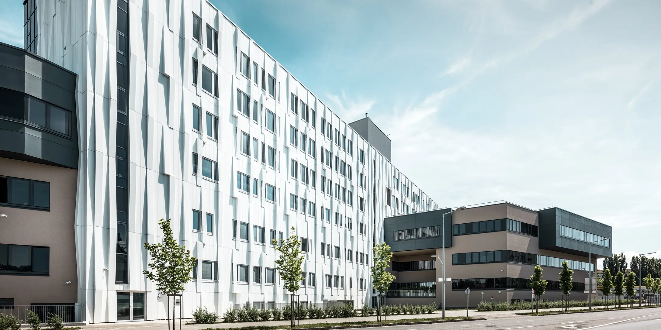 Bočný pohľad na univerzitnú kliniku v St. Pölten, Dolné Rakúsko. Robustná hliníková fasáda robí z budovy pútavý objekt v meste. V popredí je vidieť ulicu s niekoľkými stromami.