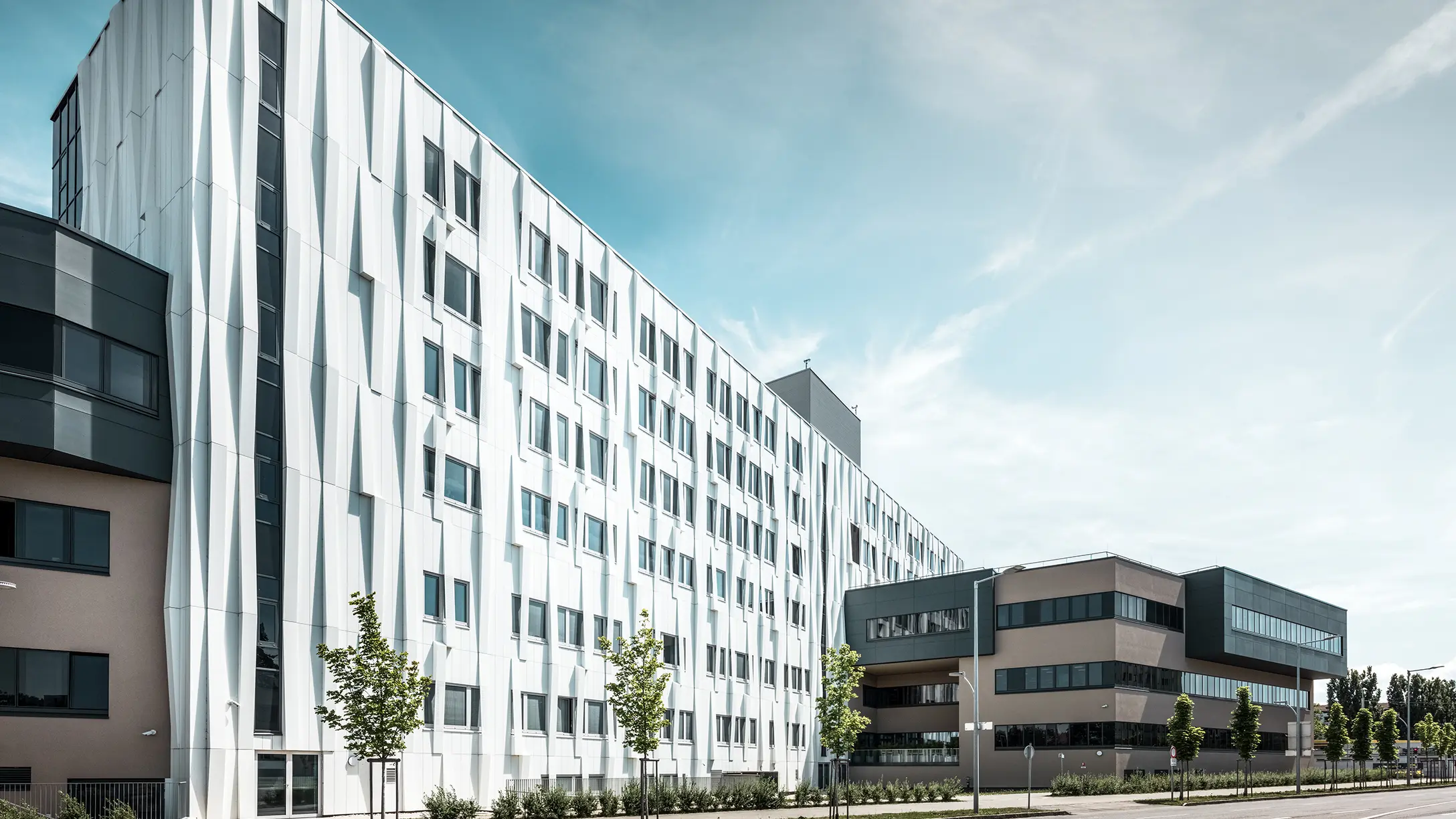 Bočný pohľad na univerzitnú kliniku v St. Pölten, Dolné Rakúsko. Robustná hliníková fasáda robí z budovy pútavý objekt v meste. V popredí je vidieť ulicu s niekoľkými stromami.
