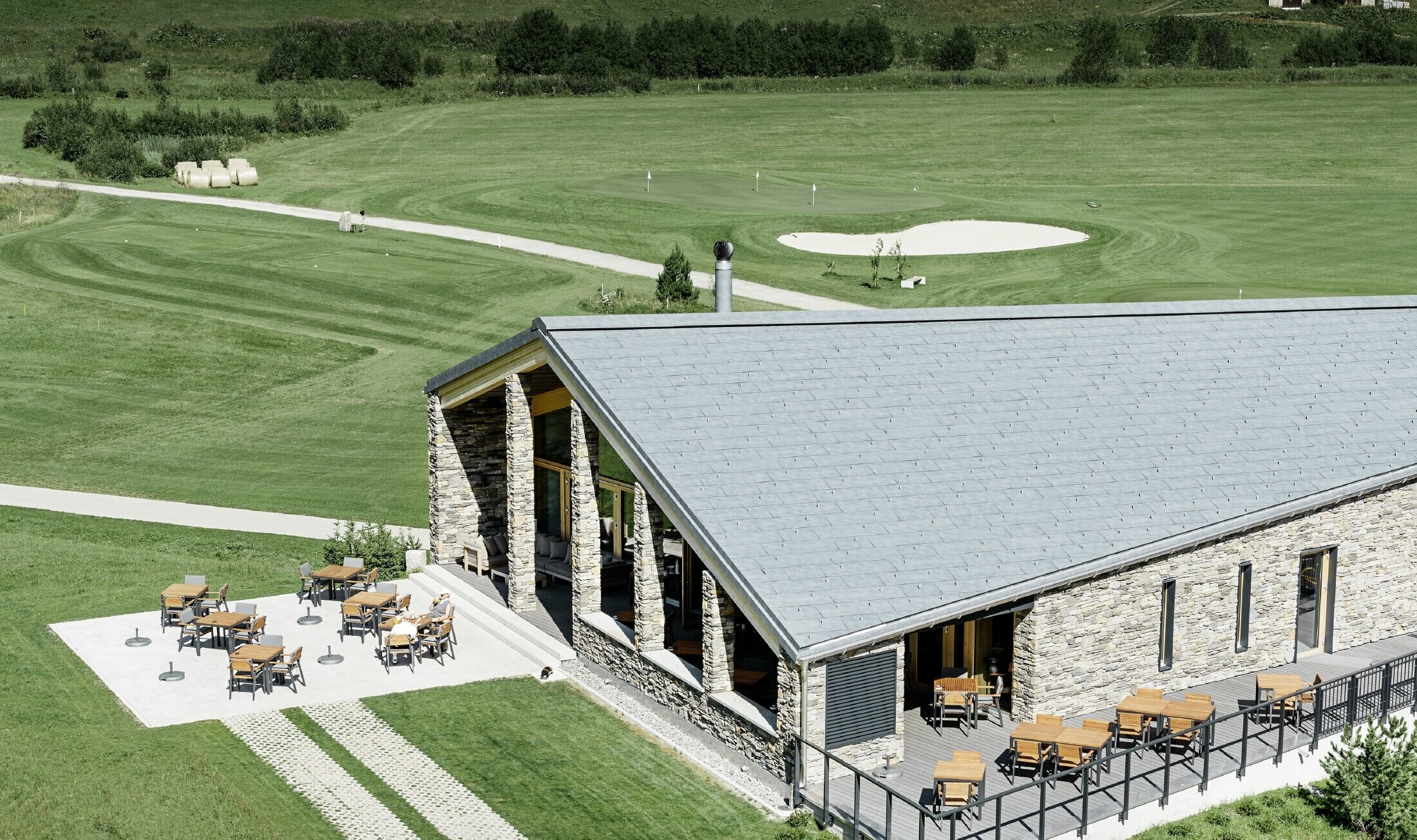 Moderná klubovňa golfového ihriska v Andermatte s kamennou fasádou a strešným panelom PREFA FX.12 z hliníka vo farbe kamenná šedá