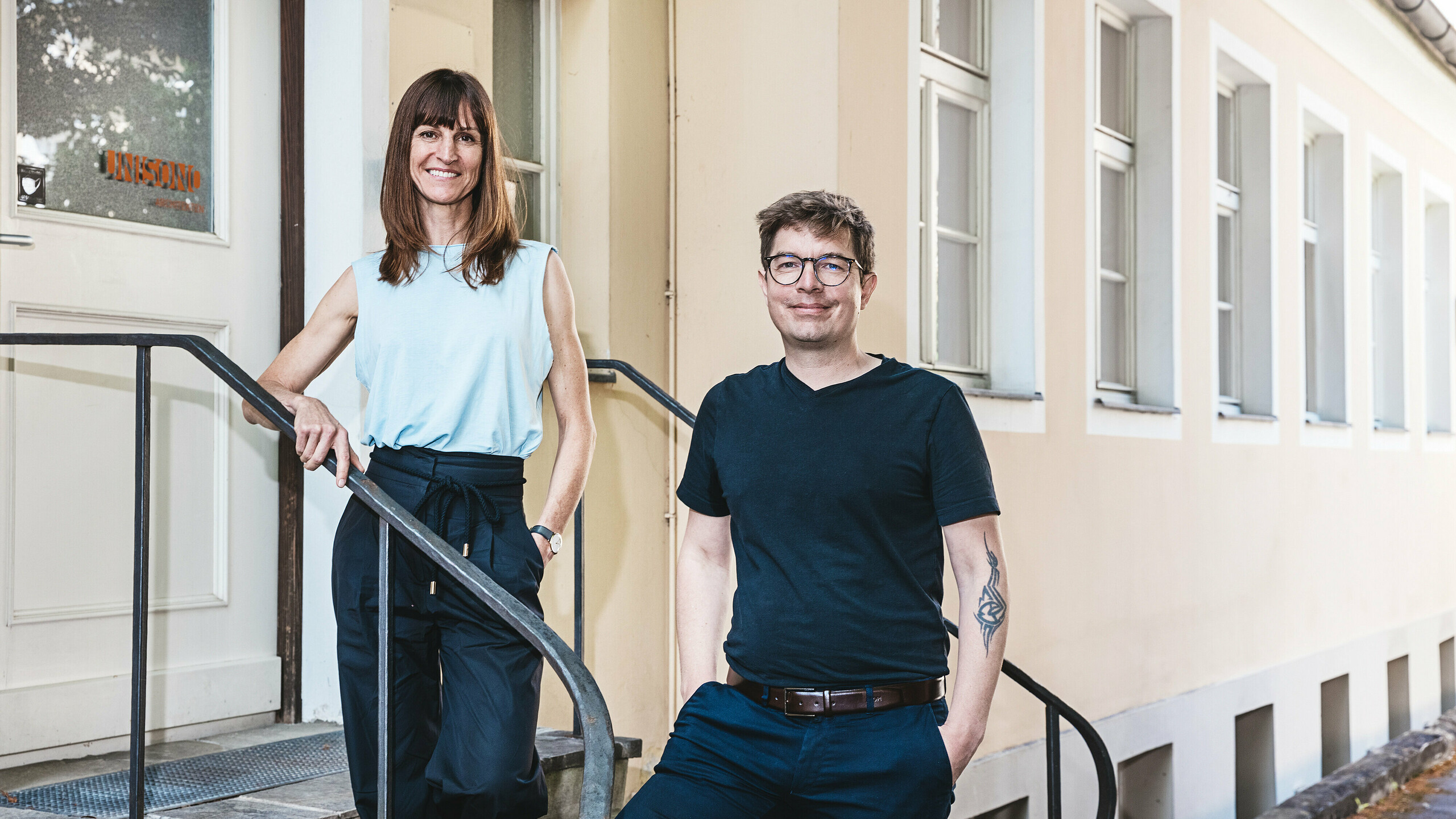 Hlavní architekti z UNISONO ARCHITEKTEN z Innsbrucku stoja na kamennom schodisku pred béžovo natretou budovou s bielymi okennými rámami. Žena má na sebe modrú blúzku bez rukávov a čierne nohavice, muž má na sebe čierne tričko a modré nohavice s hnedým opaskom. Priateľsky sa usmievajú do kamery, čo navodzuje otvorenú a prívetivú atmosféru.