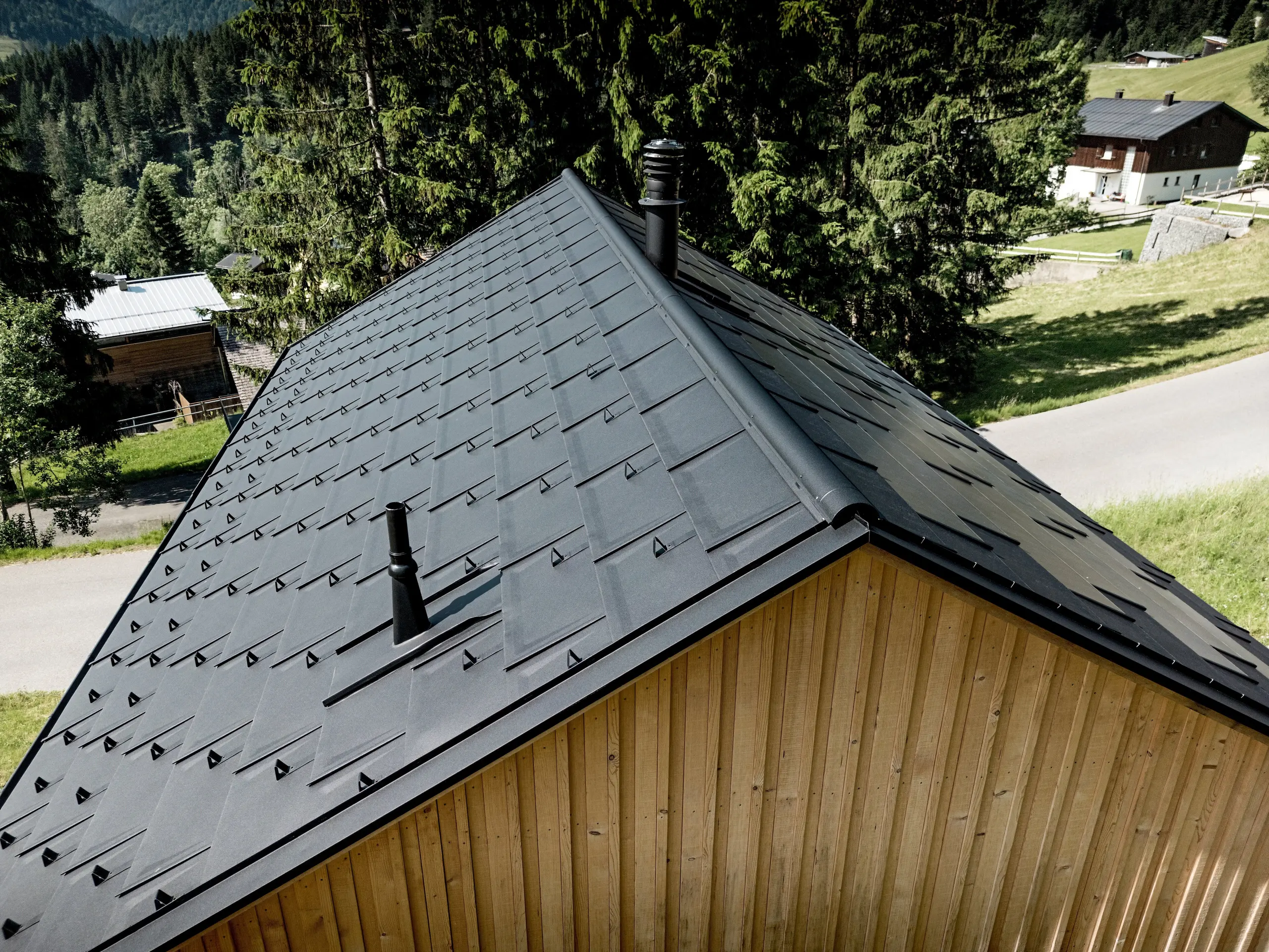 Dynamický detailný záber zachytáva výraznú strešnú plochu domu Immi na pozadí zelených horských lúk a tmavých smrekov. Strešné dosky PREFA R.16 v odtieni P.10 čierna, precízne detaily a snehové zábrany vytvárajú pôsobivý, jednotný vzhľad strechy. Ľahká a odolná hliníková krytina odoláva alpskému počasiu aj vysokému zaťaženiu snehom a chráni drevodom v Ebnite trvalo a s minimálnou údržbou.