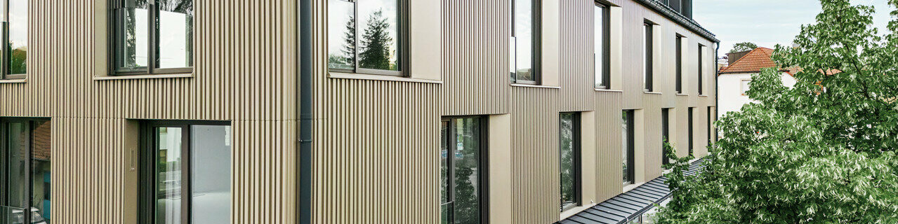 Na snímke je moderná budova vo Feldkirchene, ktorú navrhol architekt Stefan Heigl. Fasáda je z materiálu Prefalz vo farbe bronzová a šikmá strecha je pokrytá materiálom Prefalz v čiernošedej farbe. Veľké okná poskytujú vo vnútri dostatok svetla, zatiaľ čo presklená zóna na prízemí ponúka príjemný priestor na stravovanie. Kvalitný výber materiálov a elegantný dizajn dodávajú budove moderný a štýlový vzhľad.