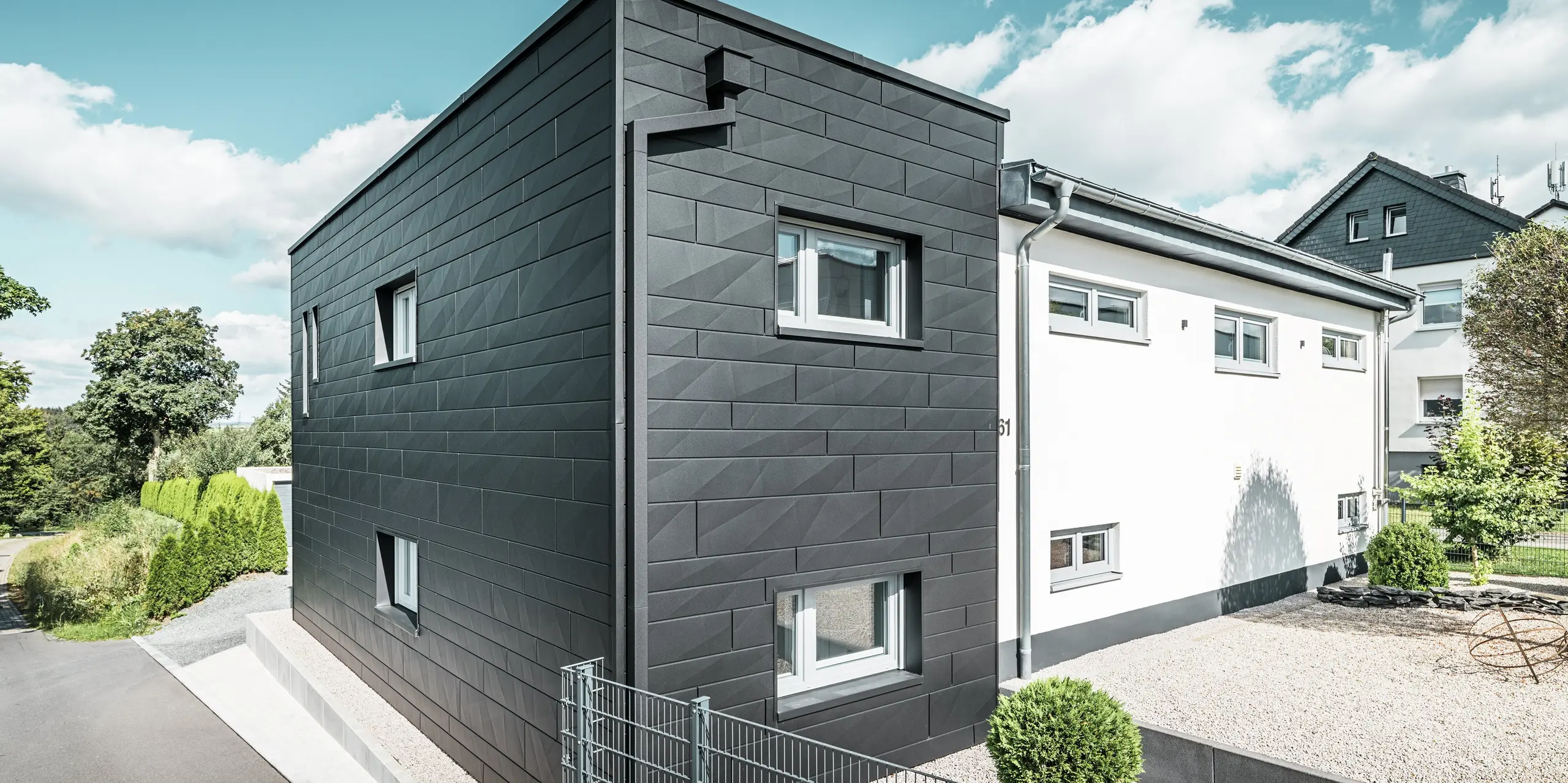 Výrazná fasáda PREFA Siding.X v čiernej farbe P.10 charakterizuje modernú architektúru zrekonštruovaného rodinného domu v Teuschnitzi. Jasne štruktúrované hliníkové panely dodávajú budove elegantnú geometrickú estetiku. Integrovaný odvodňovací systém PREFA s hranatými zvodmi sa bezproblémovo začleňuje do budovy a zaisťuje funkčné a vizuálne atraktívne riešenie. Zelená záhrada a štrková plocha pred domom dopĺňajú minimalistický dizajn a zdôrazňujú moderný charakter domu.