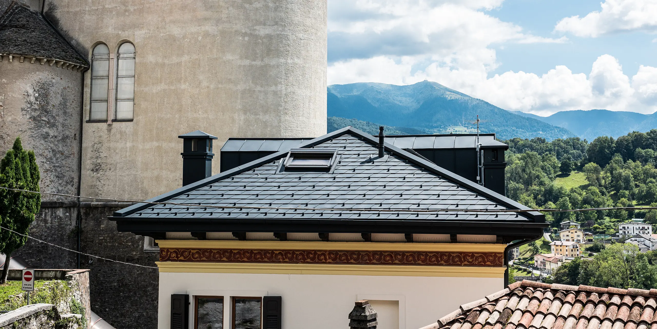 V talianskom meste Belluno sa historická a moderná architektúra spájajú v dôkladne zmodernizovanej obytnej budove, ktorej strecha je pokrytá hliníkovými šindľami PREFA antracitovej farby. Budova je v súlade s okolitou tradičnou štruktúrou a zeleným, kopcovitým pozadím, zatiaľ čo čisté línie a opláštenie komína zdôrazňujú moderný dizajn a inovatívne využitie odolných materiálov.