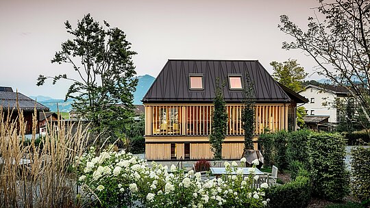 „Kúpeľný dom“ hotela Hirschen v Schwarzenbergu je obklopený starostlivo upravenou záhradou a nachádza sa priamo pred romantickým západom slnka. V popredí kvitnú biele hortenzie, okrasné trávy a kríky, ktoré rámujú otvorený vonkajší priestor s posedením a ohniskom. V centre stojí moderná budova s nápadnou strechou PREFA vo farbe P.10 orieškovohnedá. Jej presne spracované plechy sú šikmé a dodávajú streche dynamický efekt. Fasáda z vertikálnych drevených lamiel po celej výške poschodia spája moderný dizajn s regionálnym štýlom stavby. V pozadí sa rysujú mierne kopce a hory Bregenzerwaldu – harmonické spojenie prírody, architektúry a tradície.