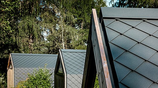 Niekoľko minimalistických minidomov typu A-Frame v areáli Secret Koi Garden sa harmonicky radí na okraji lesa pri jazere Natterer See. Strešné plochy sú pokryté malými hliníkovými šablónami PREFA v odtieni P.10 antracitová, ktoré vytvárajú výraznú štruktúru pripomínajúcu rybiu kožu. Matný povrch pôsobí pokojne a elegantne, zatiaľ čo odolný a poveternostne odolný hliník PREFA ponúka optimálnu ochranu v alpskom prostredí a dokonale spája dizajn s prírodou.