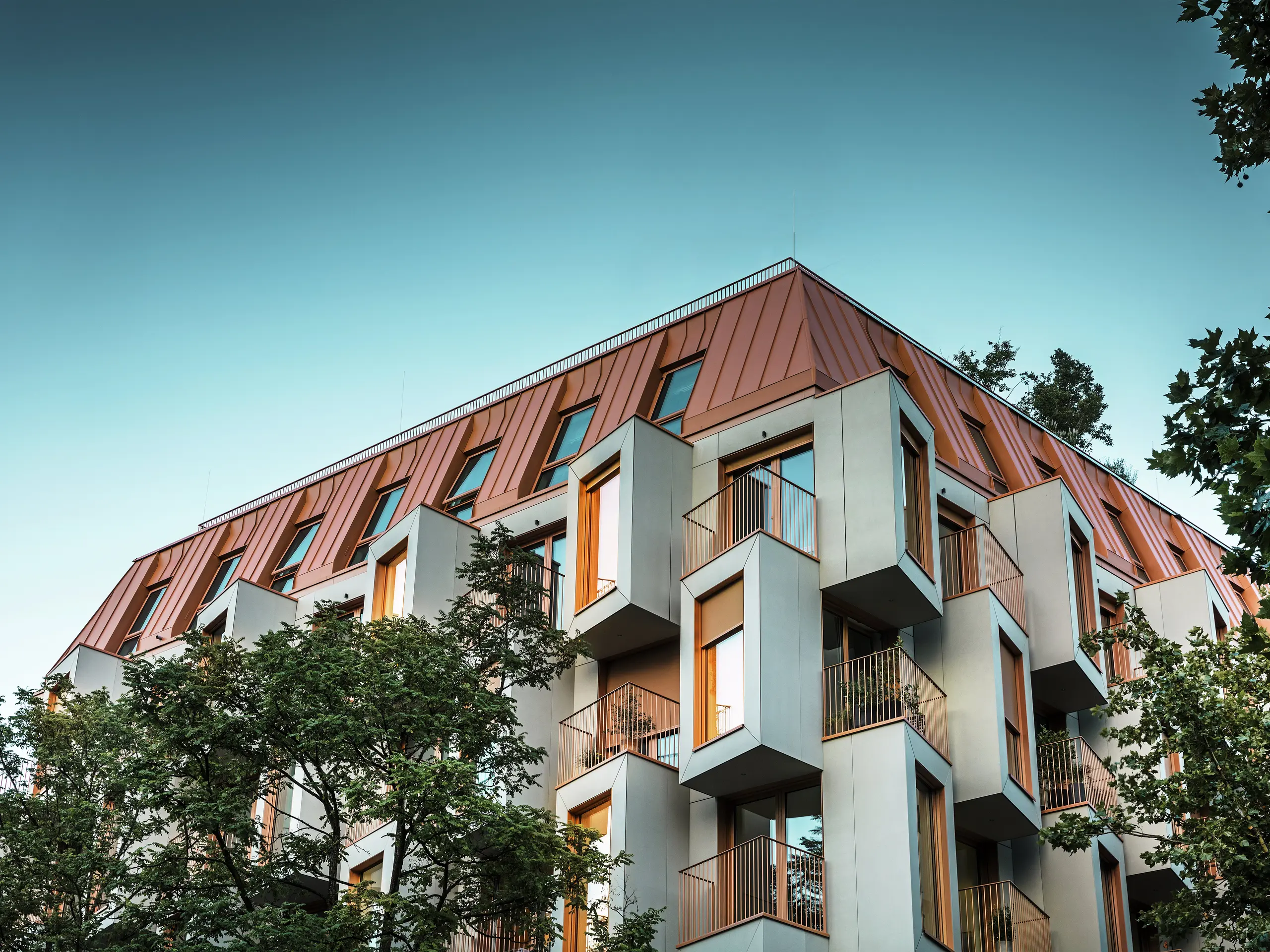 Architektonicky výnimočná fasáda obytného projektu Van B v Mníchove s výraznými arkierovými oknami a hliníkovou strechou PREFALZ v špeciálnej farbe IGP Orange HDP. Pri pohľade zdola za súmraku sa v oknách objavujú svetlá. Moderné riešenie strechy spája maximálnu odolnosť voči poveternostným vplyvom s elegantným vzhľadom a v mestskom priestore vytvára farebné akcenty. Súhra sklovláknobetónu, medenooranžových okenných rámov a hliníkovej strechy vytvára dynamický, odolný a udržateľný plášť budovy - ideálny pre výstavbu orientovanú na budúcnosť.