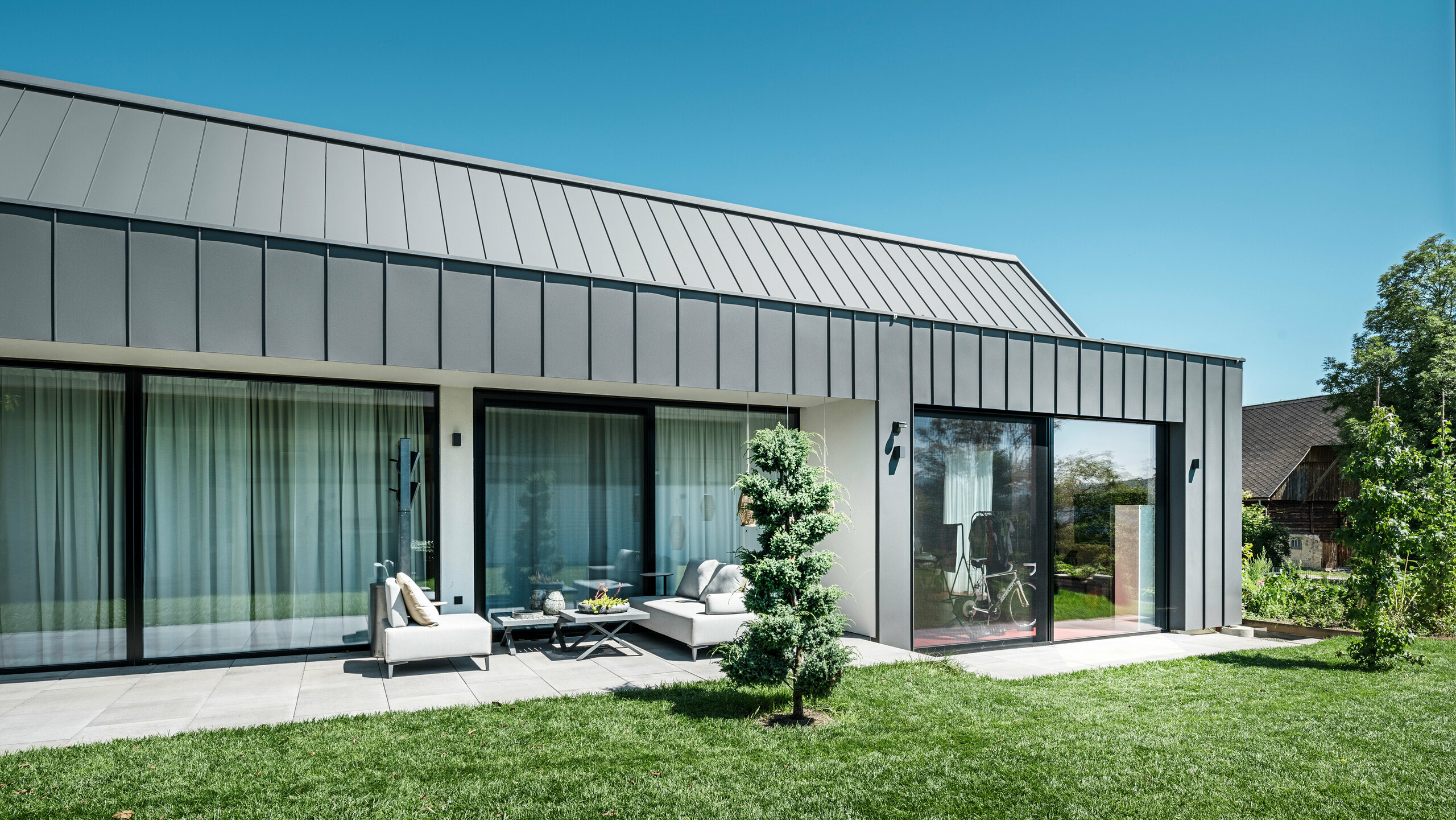 Uvoľnená záhradná terasa rodinného domu v rakúskom Pogieri s elegantným strešným a fasádnym obkladom PREFALZ vo farbe P.10 tmavošedá od firmy PREFA. Veľké presklené čelá otvárajú obytný priestor prírode a umožňujú plynulý prechod medzi vnútornými a vonkajšími priestormi. Moderný dizajn sa stretáva s funkčnosťou, pričom zelené okolie a starostlivo upravený trávnik umocňujú pokojnú atmosféru miesta
