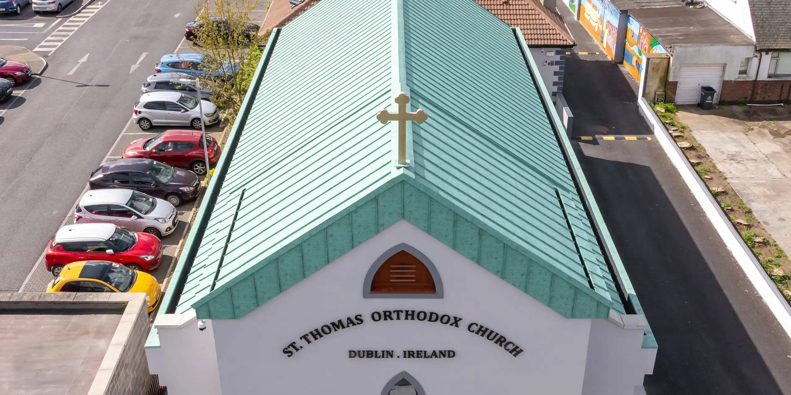 Drohnenaufnahme der Frontansicht der St. Thomas Orthodox Church in Dublin mit ihrem markanten PREFALZ Aluminium-Stehfalzdach in P.10 Patinagrün. Das weithin sichtbare Kreuz am Giebel, die eleganten Rundbogenfenster und die klar gegliederte Fassade verleihen dem Sakralbau eine unverwechselbare Präsenz. Die großflächige Dachlandschaft mit präzisen Stehfalzlinien und der charakteristischen Patinaoptik erinnert an traditionelle Kupferdächer und setzt einen starken architektonischen Akzent im urbanen Umfeld. Das hochwertige PREFA Aluminium überzeugt durch Korrosionsbeständigkeit, geringes Gewicht, Langlebigkeit und dauerhafte Farbqualität – ideal für anspruchsvolle Architektur und nachhaltige Dachlösungen.